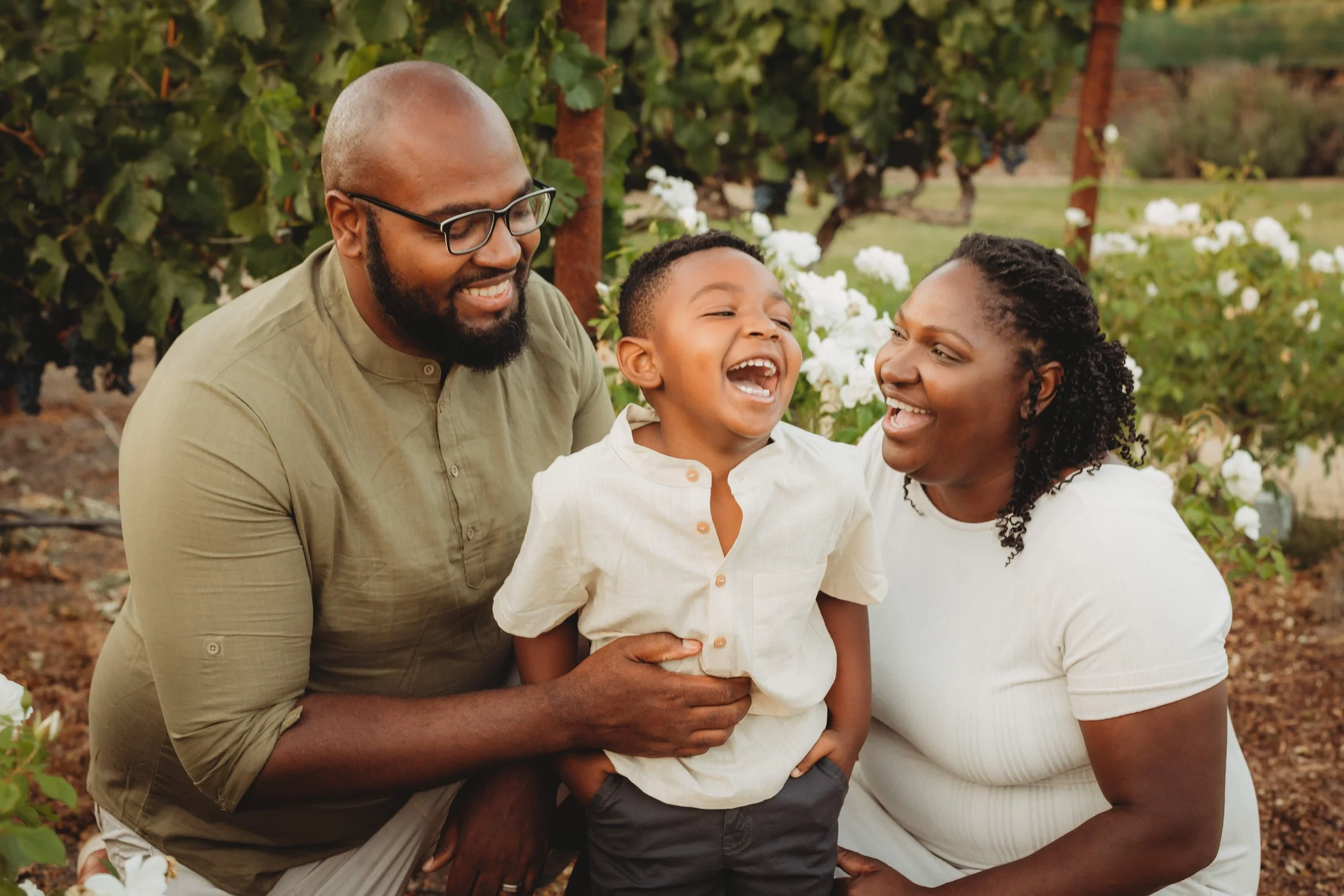 A happy family, including a man, woman, and young boy, laughing together outdoors in a garden or park with greenery and white flowers.
