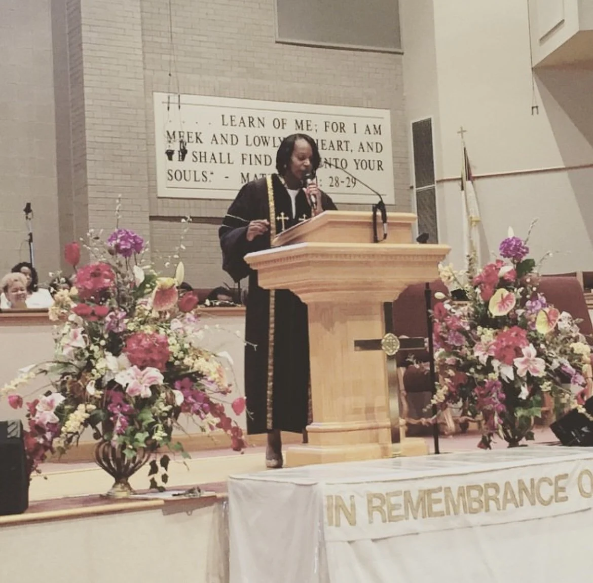 A woman dressed in a black robe with gold crosses standing at a wooden podium speaking into a microphone, surrounded by colorful flower arrangements, in a church setting with a large banner behind her reading scripture from Matthew 11:28-29.