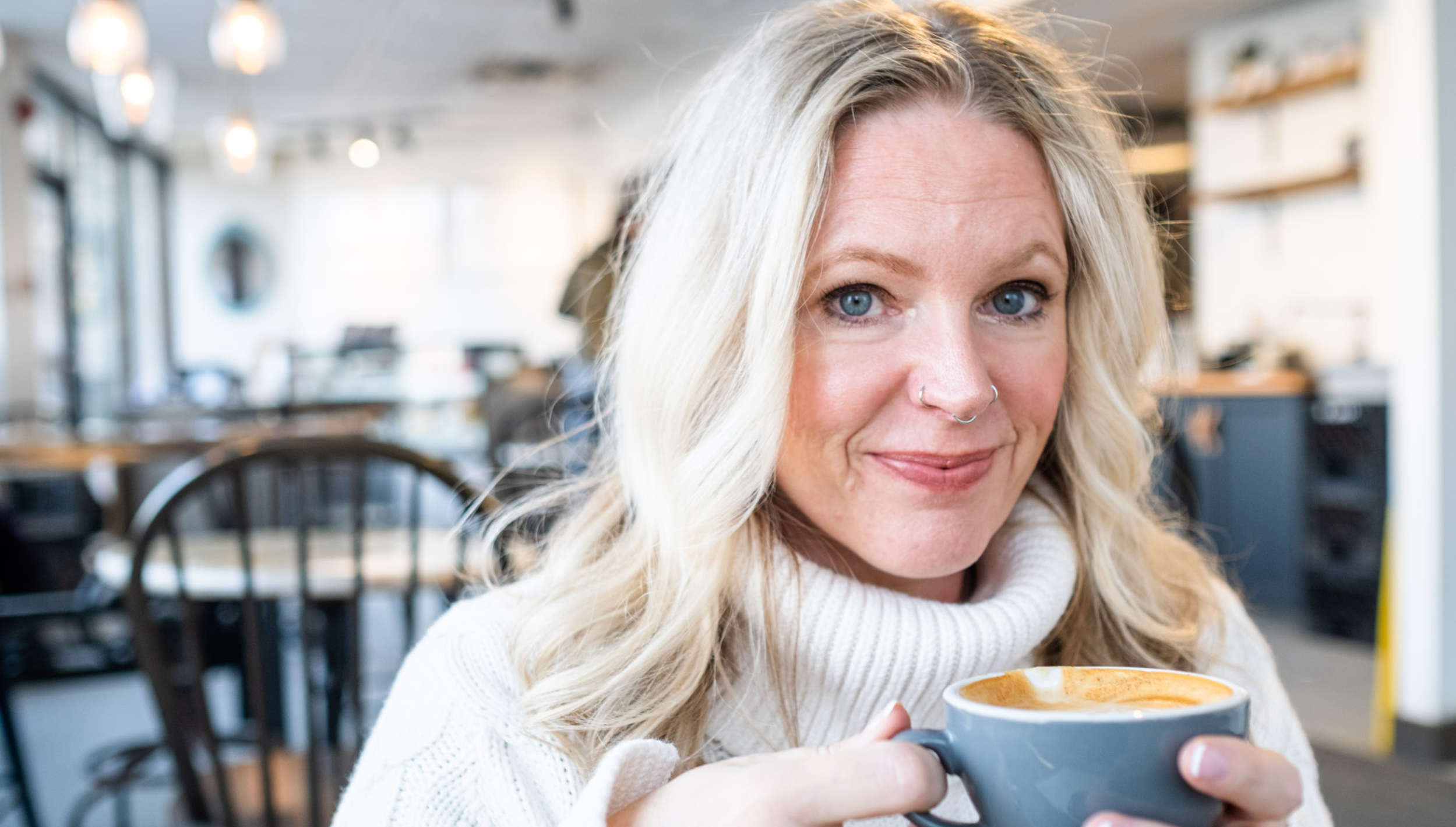 A woman with blonde hair and blue eyes smiling in a cafe, holding a cup of coffee with a latte art. She is wearing a white turtleneck sweater.