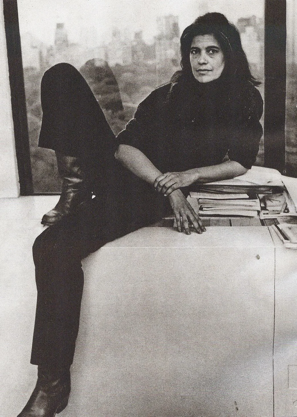 Essayist Susan Sontag relaxes on a window sill that overlooks what appears to be New York City. She wears dark clothes and boots with heels.