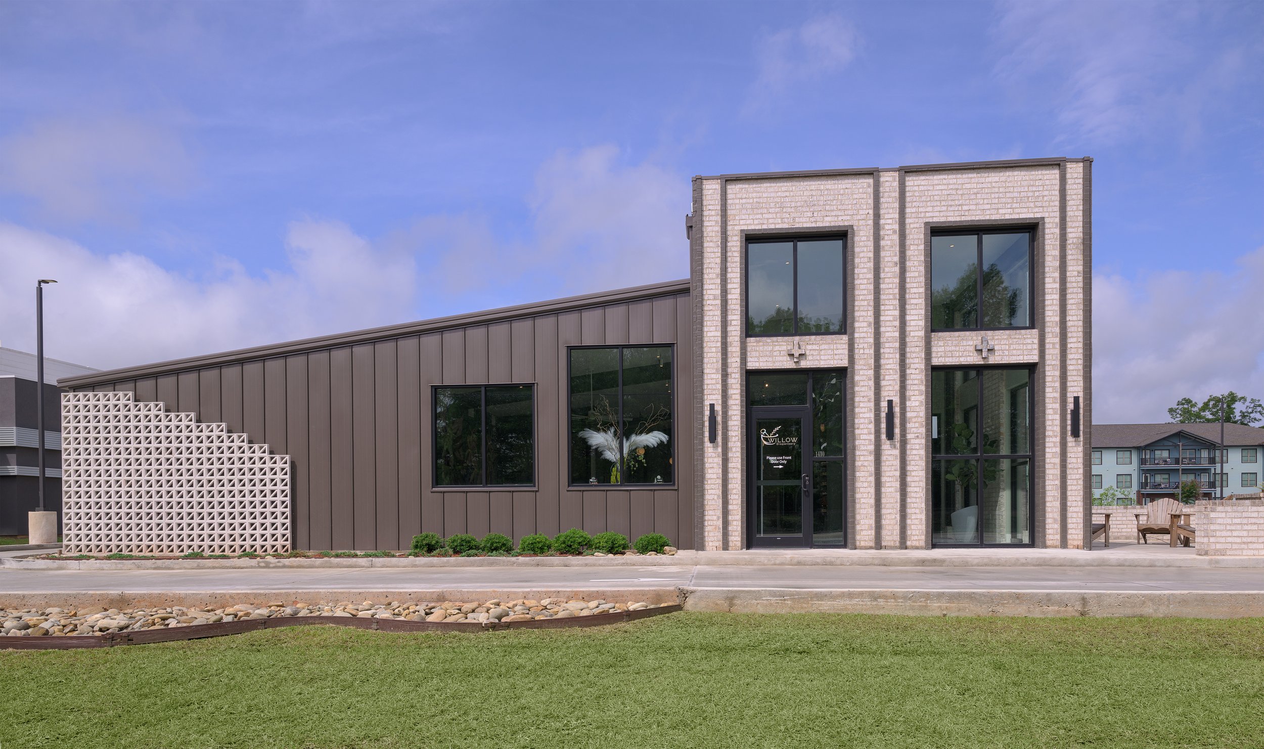 Modern building dispensary called Willow Pharmacy with large windows, beige brick facade, and gray siding, with a small front lawn and walkway, under a blue sky with some clouds.