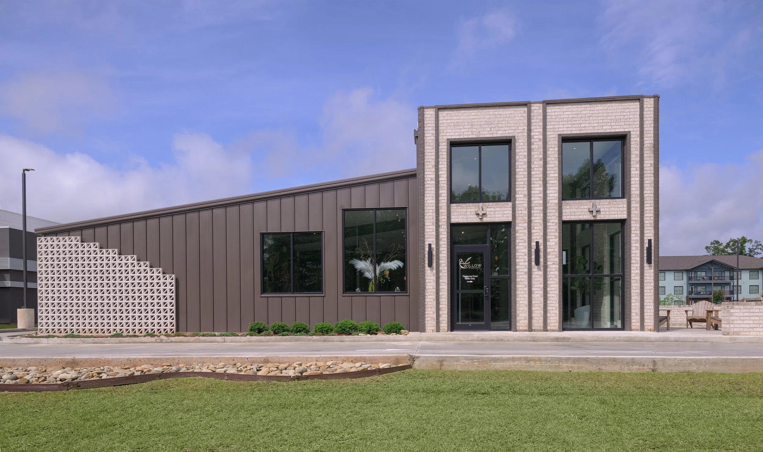 Modern building dispensary called Willow Pharmacy with large windows, beige brick facade, and gray siding, with a small front lawn and walkway, under a blue sky with some clouds.