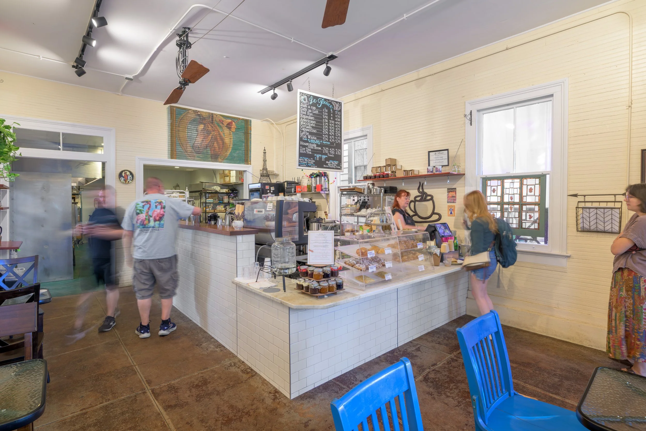 Interior of a café with a counter selling baked goods, menu board hanging above, and customers waiting and sitting at blue chairs.
