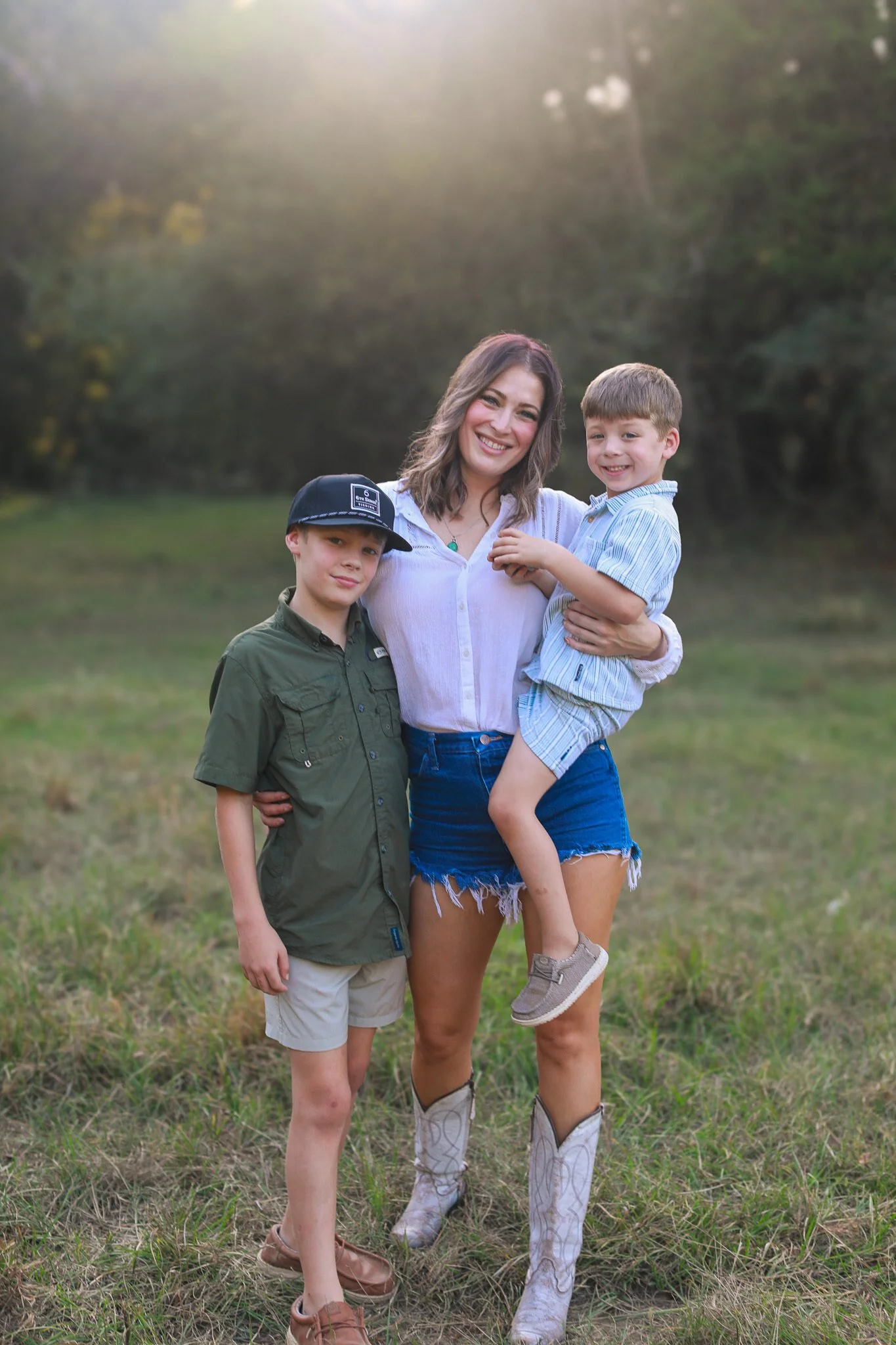 A woman and two young boys outdoors in a grassy field, smiling at the camera, during sunset or sunrise.