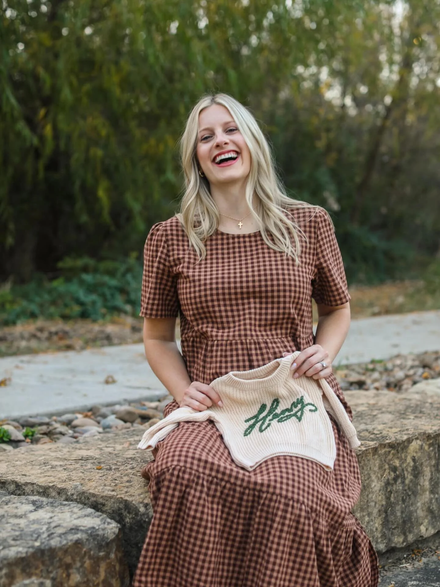 A young woman with blonde hair sits on a large stone outdoors, smiling and holding a knitted sweater that says 'Hope' with a heart. She is wearing a brown and black checkered dress, and there are trees and foliage in the background.
