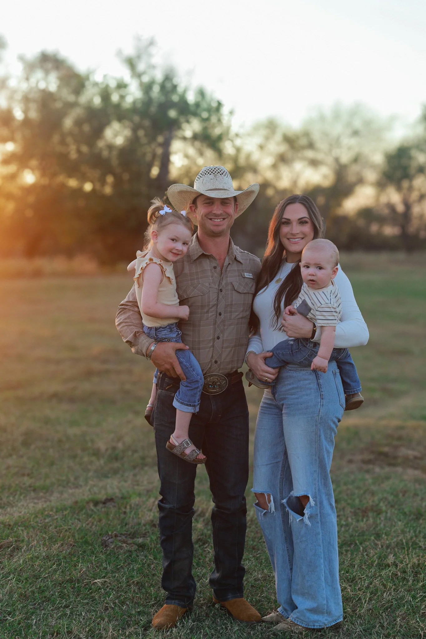 A family of four standing outdoors at sunset, with trees in the background. The father wears a cowboy hat and plaid shirt, holding a young girl. The mother, in ripped jeans and a white top, holds a baby boy.