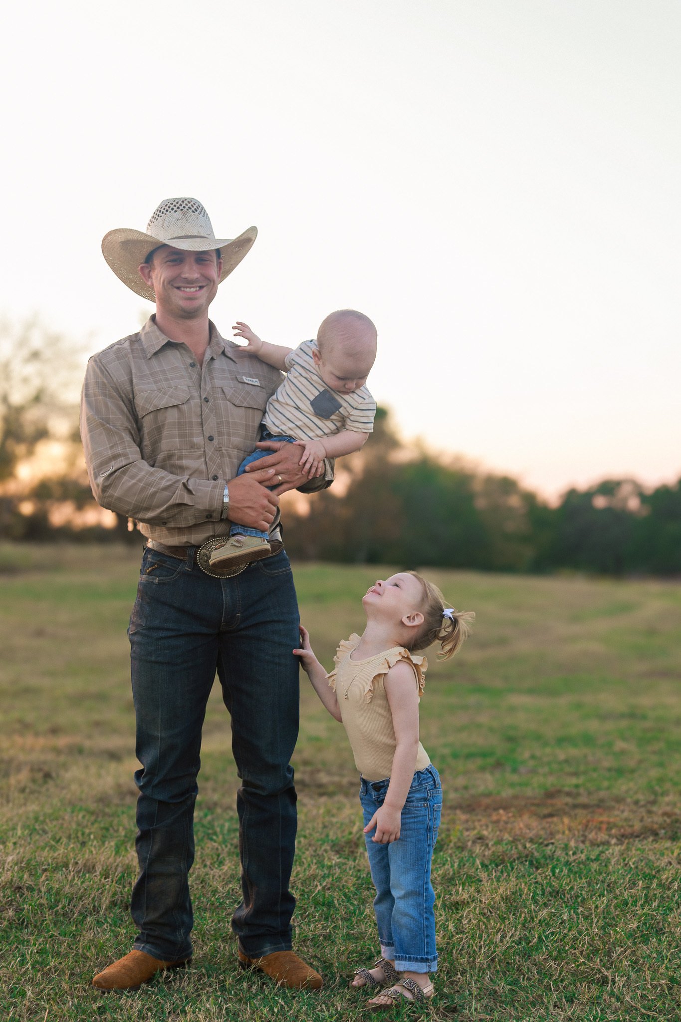 A man wearing a cowboy hat is smiling while holding a young boy, and a girl looking up at them is touching his leg, all outdoors during sunset.