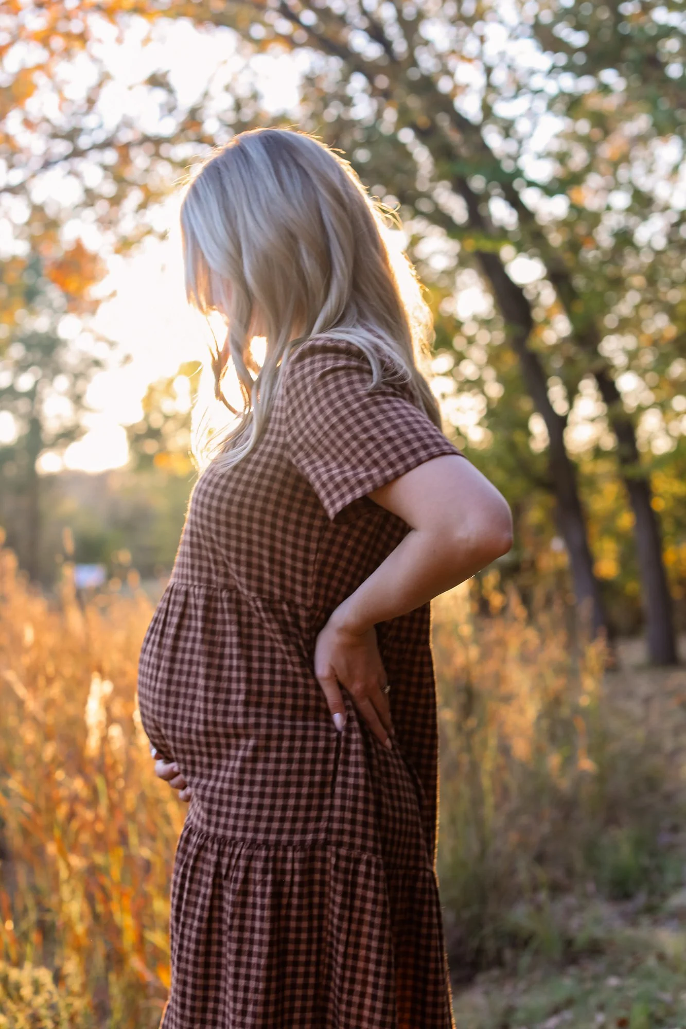 A pregnant woman in a checkered dress stands outdoors during golden hour, with trees and sunlight in the background.