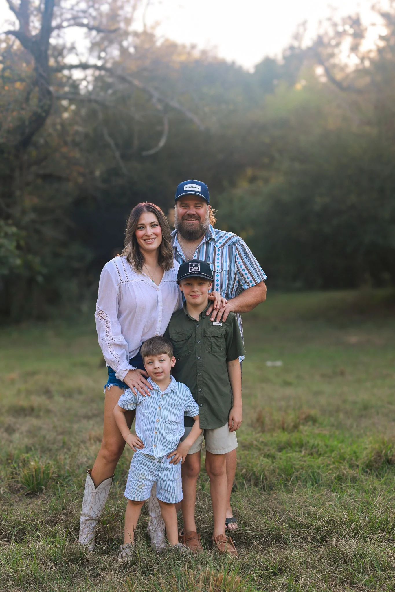 A family of five posing outdoors in a grassy field with trees in the background, during sunset. The father in a striped shirt and baseball cap, the mother in a white blouse and denim shorts, and three children, one in a plaid shirt and cowboy boots, 