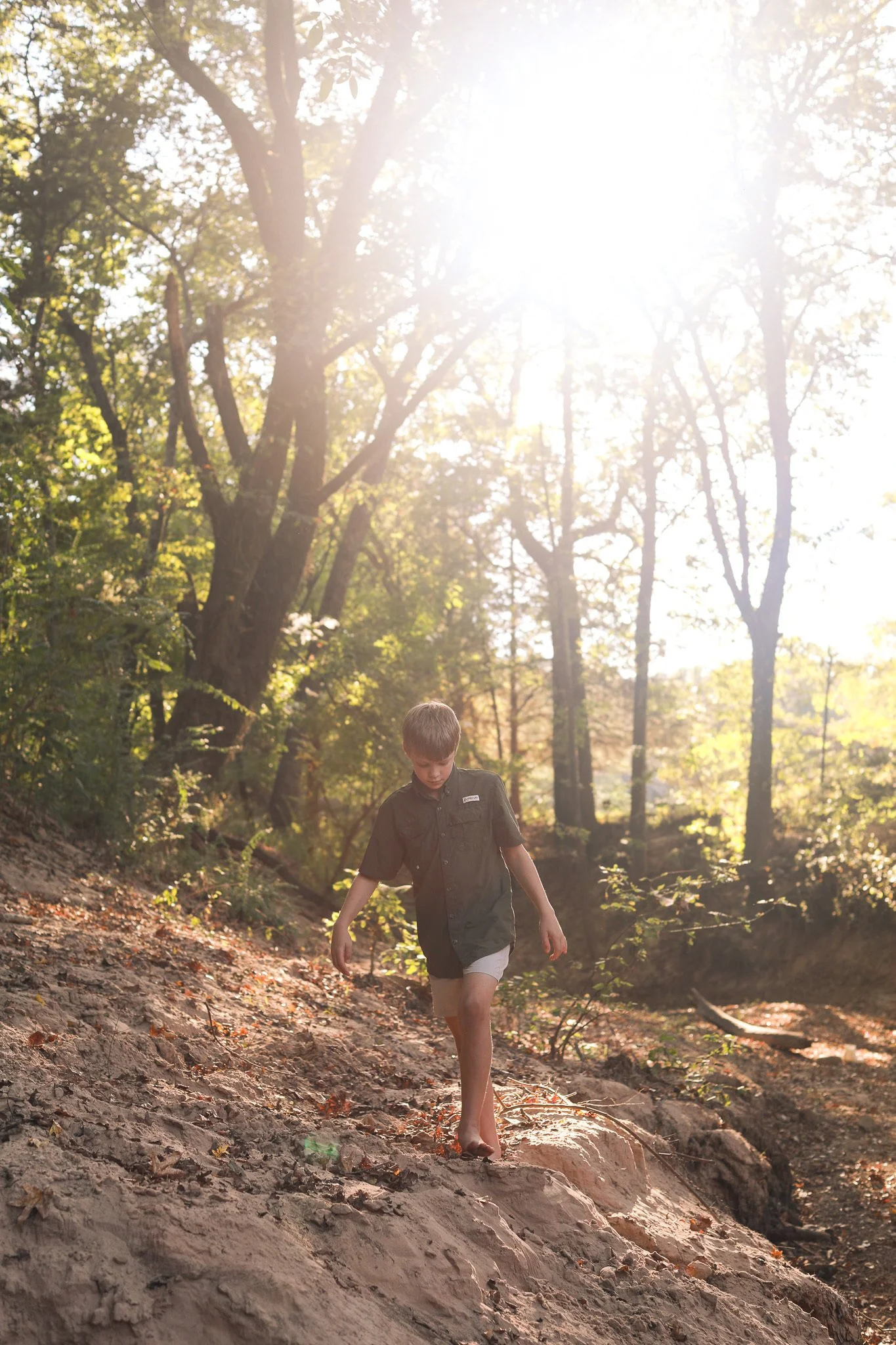 A young boy walking barefoot on a dirt trail in a wooded area during late afternoon sunlight.