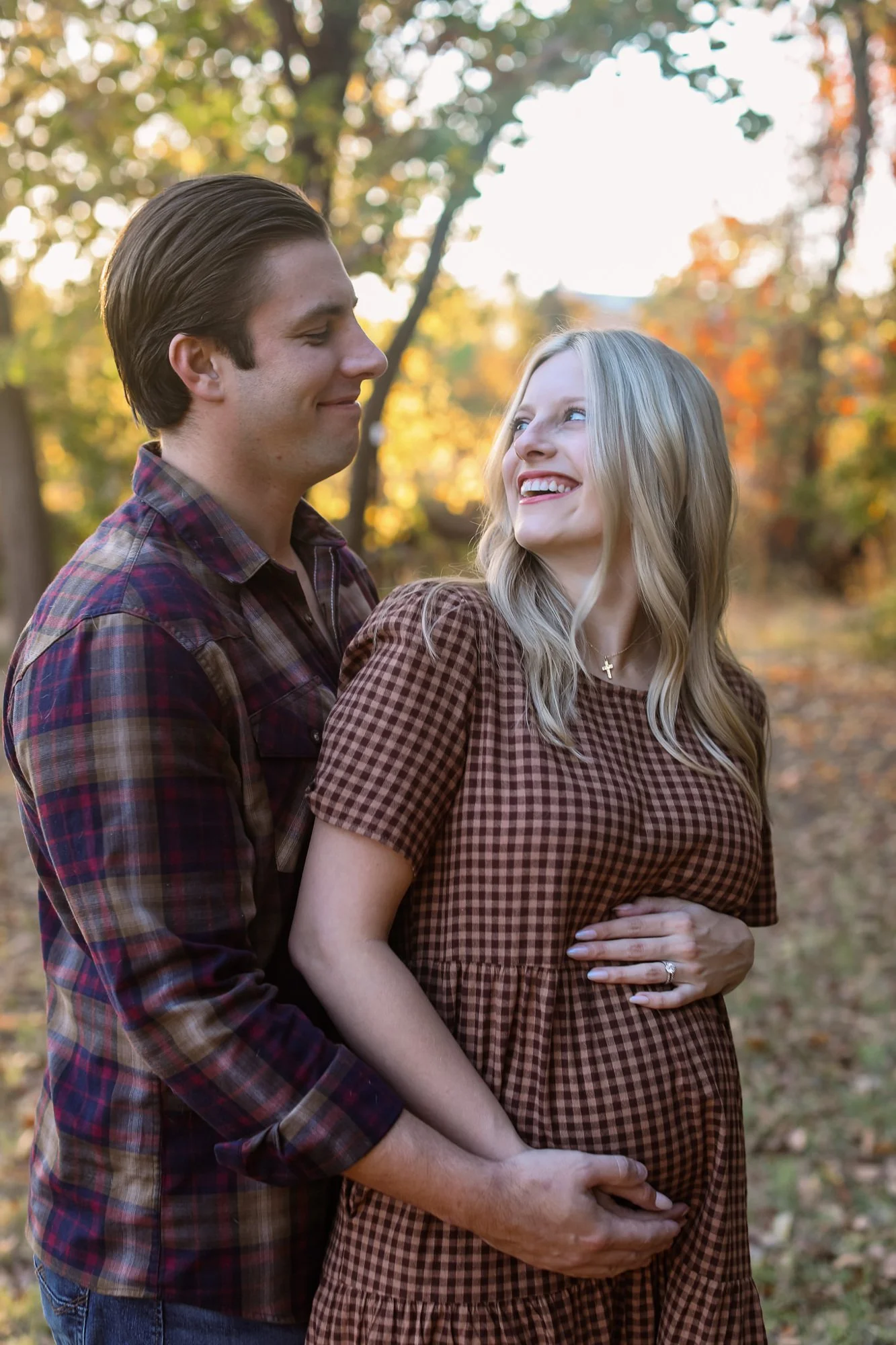 A happy couple, with the man holding the pregnant woman's belly, standing outdoors in a park during autumn, surrounded by colorful fall foliage.