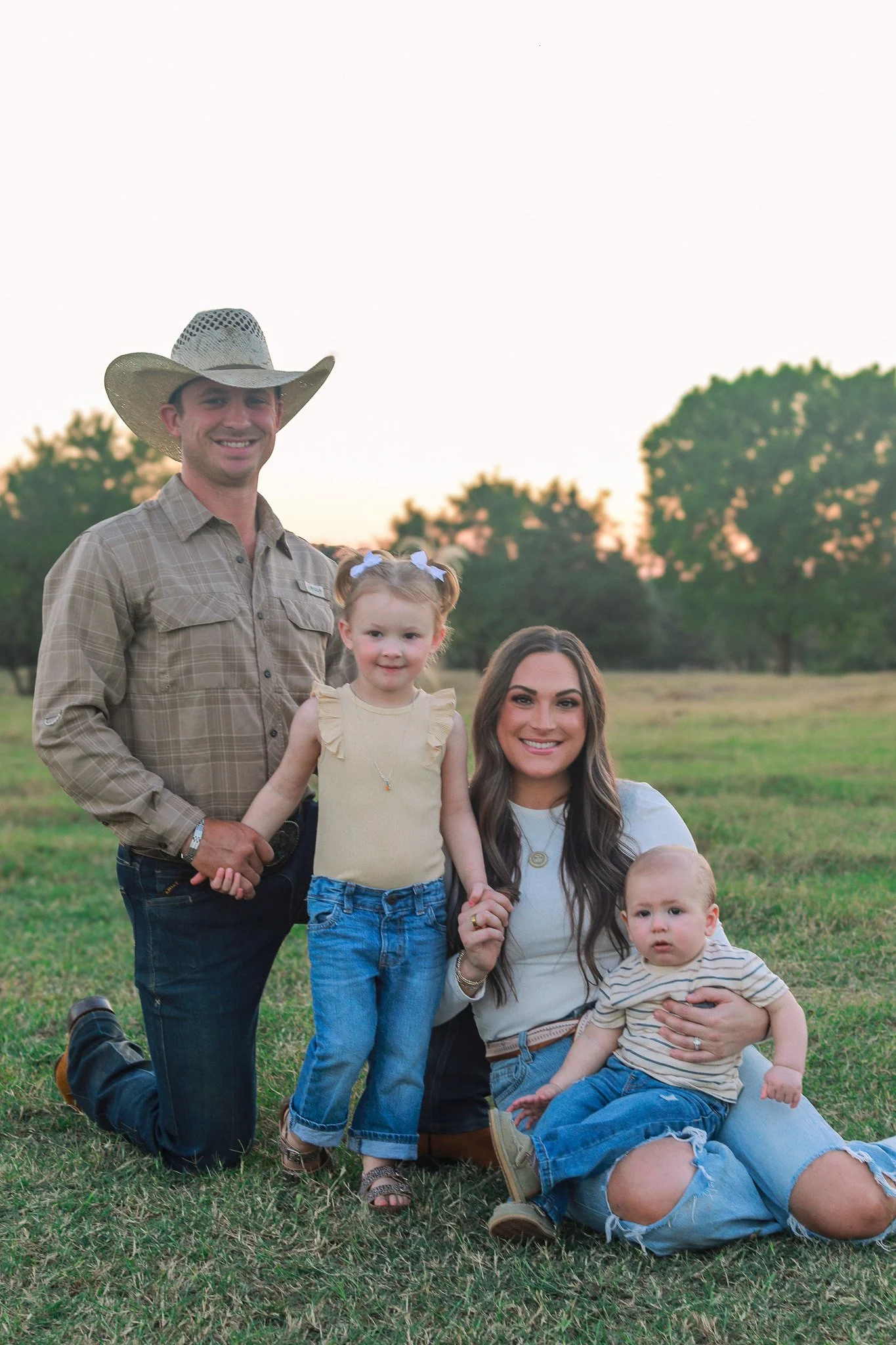 A family of four outdoors on a grassy field at sunset, with trees in the background. The father, wearing a cowboy hat and a plaid shirt, is kneeling on one knee. The mother, with long brown hair, is sitting on the ground holding a young child and smi