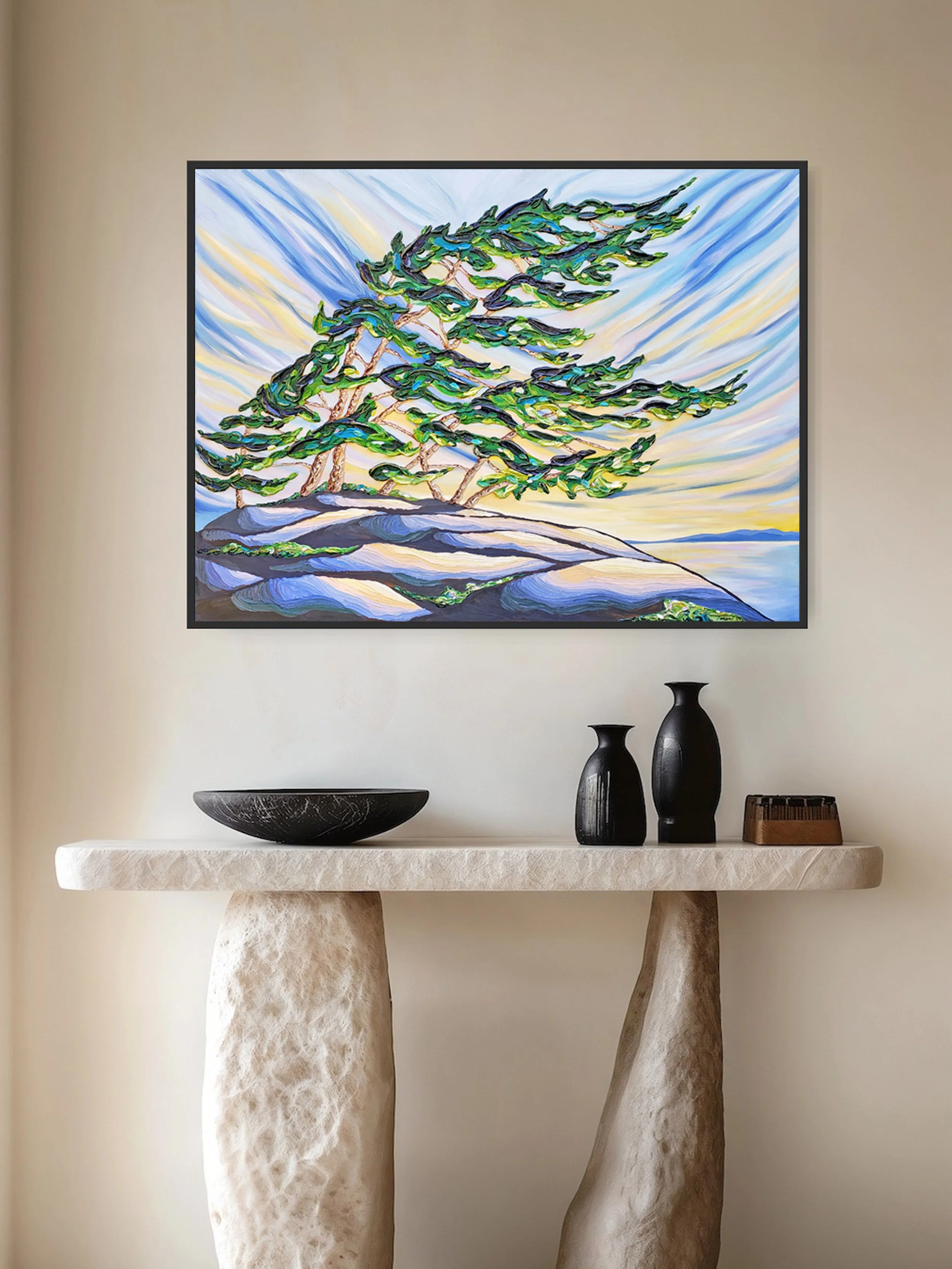 A colorful painting of windswept trees on a rocky shoreline hangs above a modern console table with black vases and a decorative bowl.