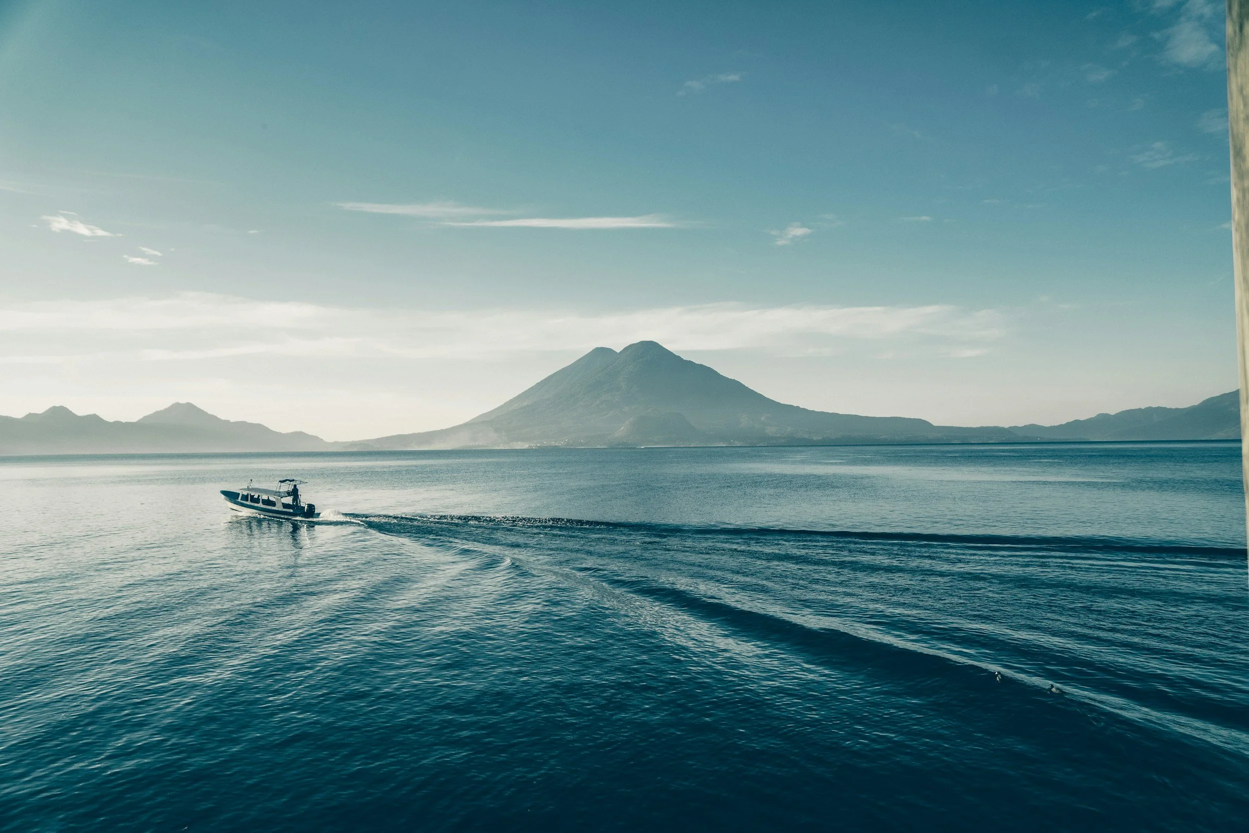 A boat sailing on a calm body of water with lush green mountain and volcano in the background under a partly cloudy sky.