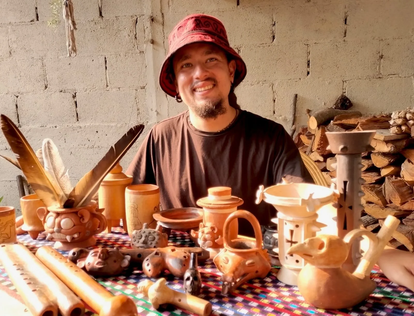 A man wearing a red hat smiling at a table displaying various pottery items and wood carvings, with a background of a brick wall and stacked firewood.