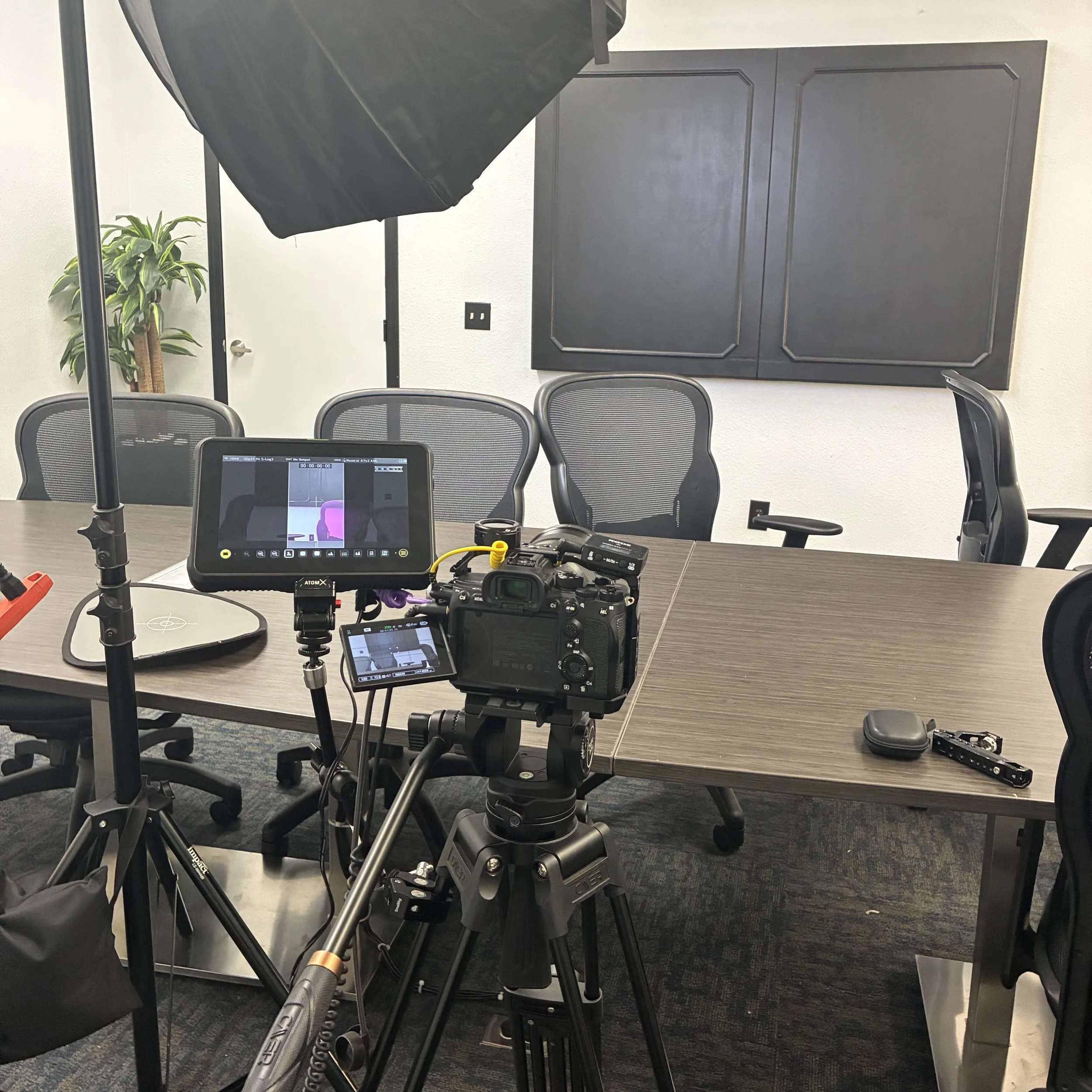 A camera setup on a tripod with an external monitor is positioned in a conference room. The room has a long table, several office chairs, a large black wall cabinet, and a potted plant in the background. Equipment on the table includes a small case and a filming accessory.