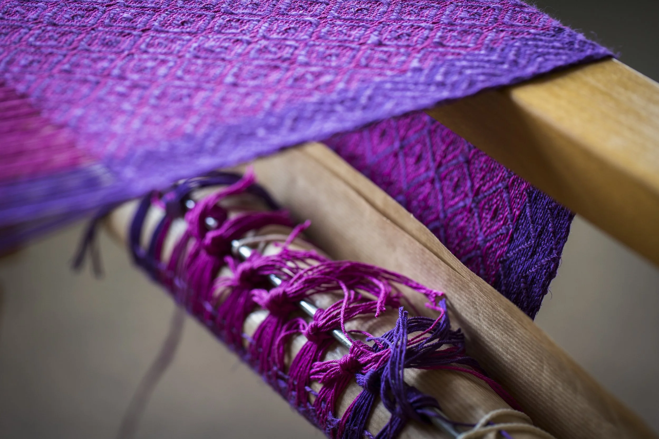 Detailaufnahme eines Webstuhls mit bunten, miteinander verflochtenen Garnen und einer gewebten Textile mit lila, pinken und violetten Mustern.