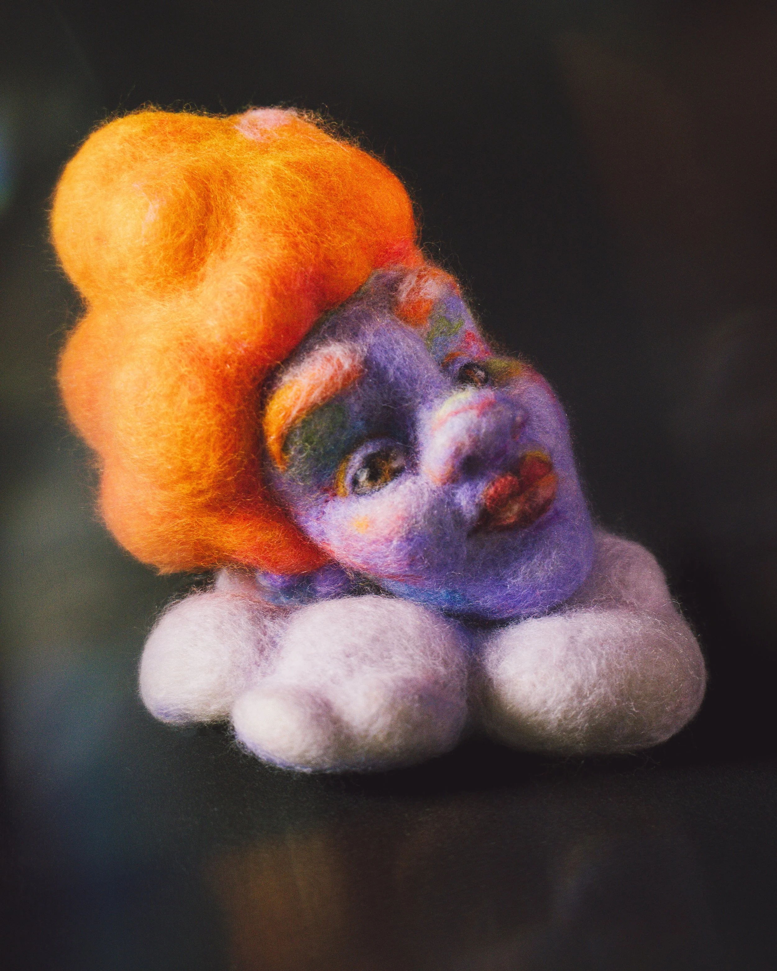 Needle felted sculpture of a lilac-skinned person with a somewhat edwardian hairstyle in bright orange. Her face is leaning slightly on a light felted cloud. All photographed with dreamy light against a dark background.