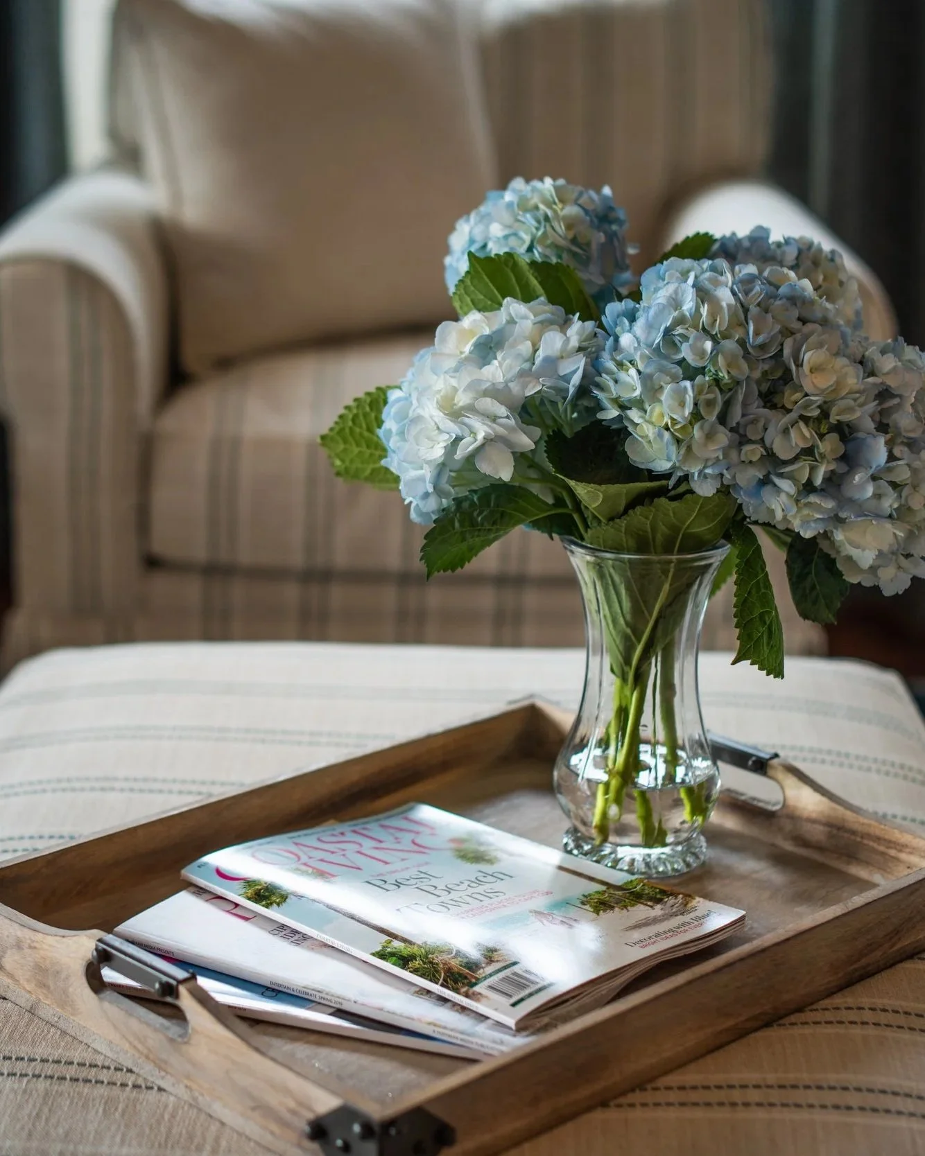 A clear glass vase with light blue and white hydrangea flowers and green leaves on a wooden tray. There are magazines underneath the tray.