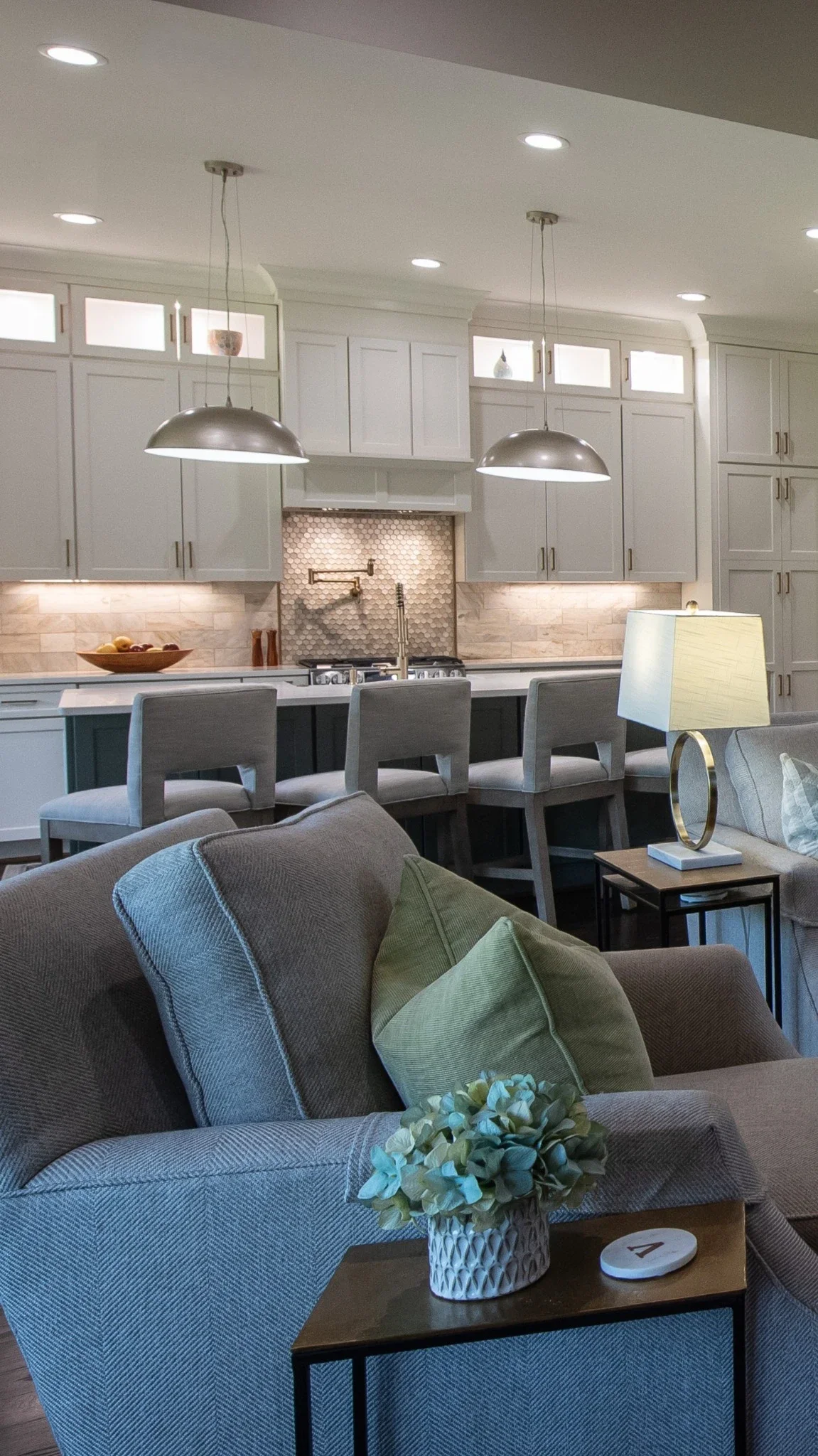 Living room with gray sofa, green throw pillows, a side table with a plant, and a kitchen with white cabinets, pendant lights, and a marble backsplash.