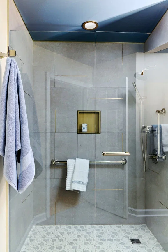 A walk-in shower with glass doors, gray tiled walls, a small built-in shelf, and a white towel hanging on a horizontal grab bar.