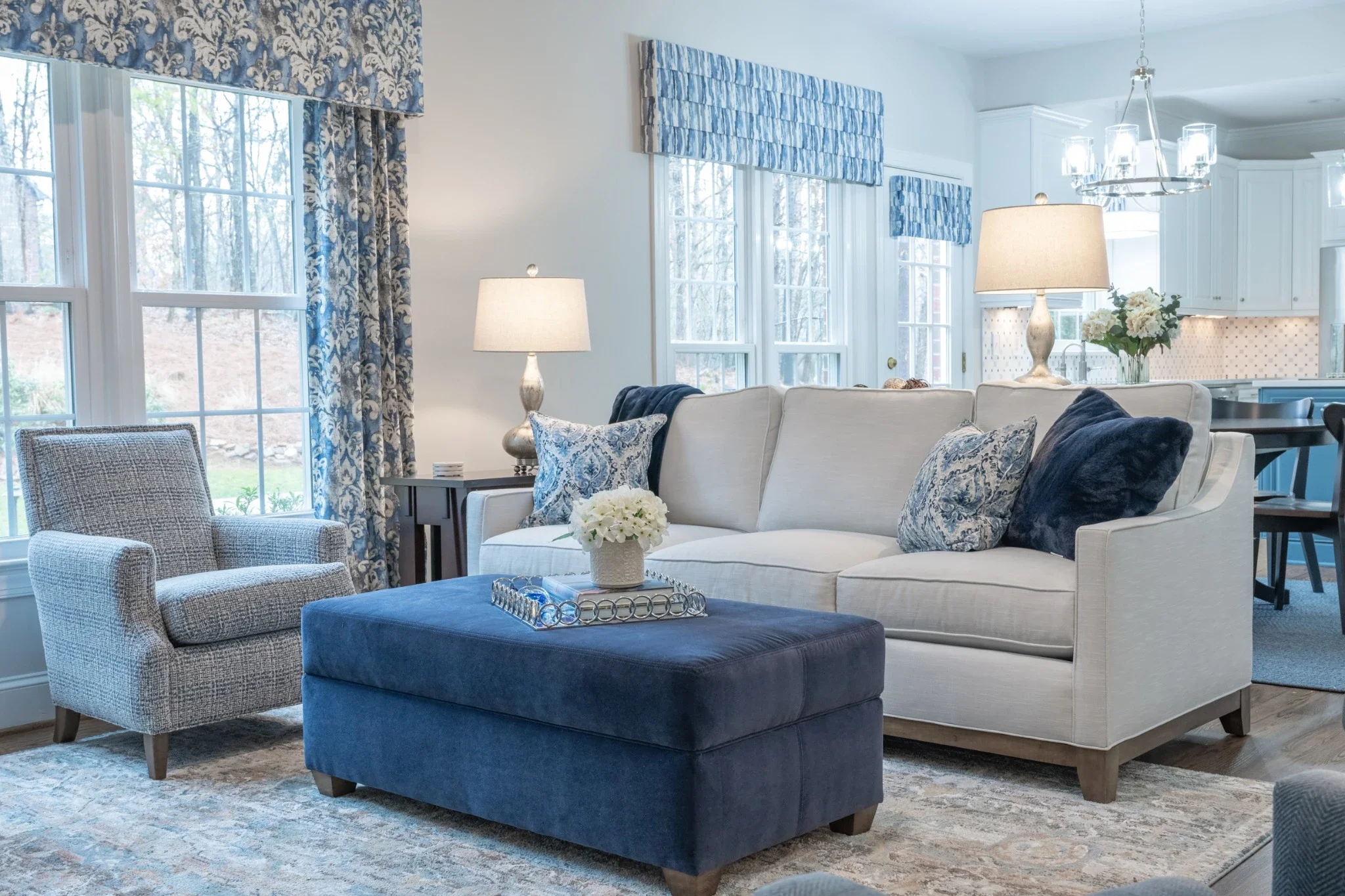 Living room with a white sofa, blue ottoman, gray armchair, lamps, curtains, and windows, with a view of the outdoors.