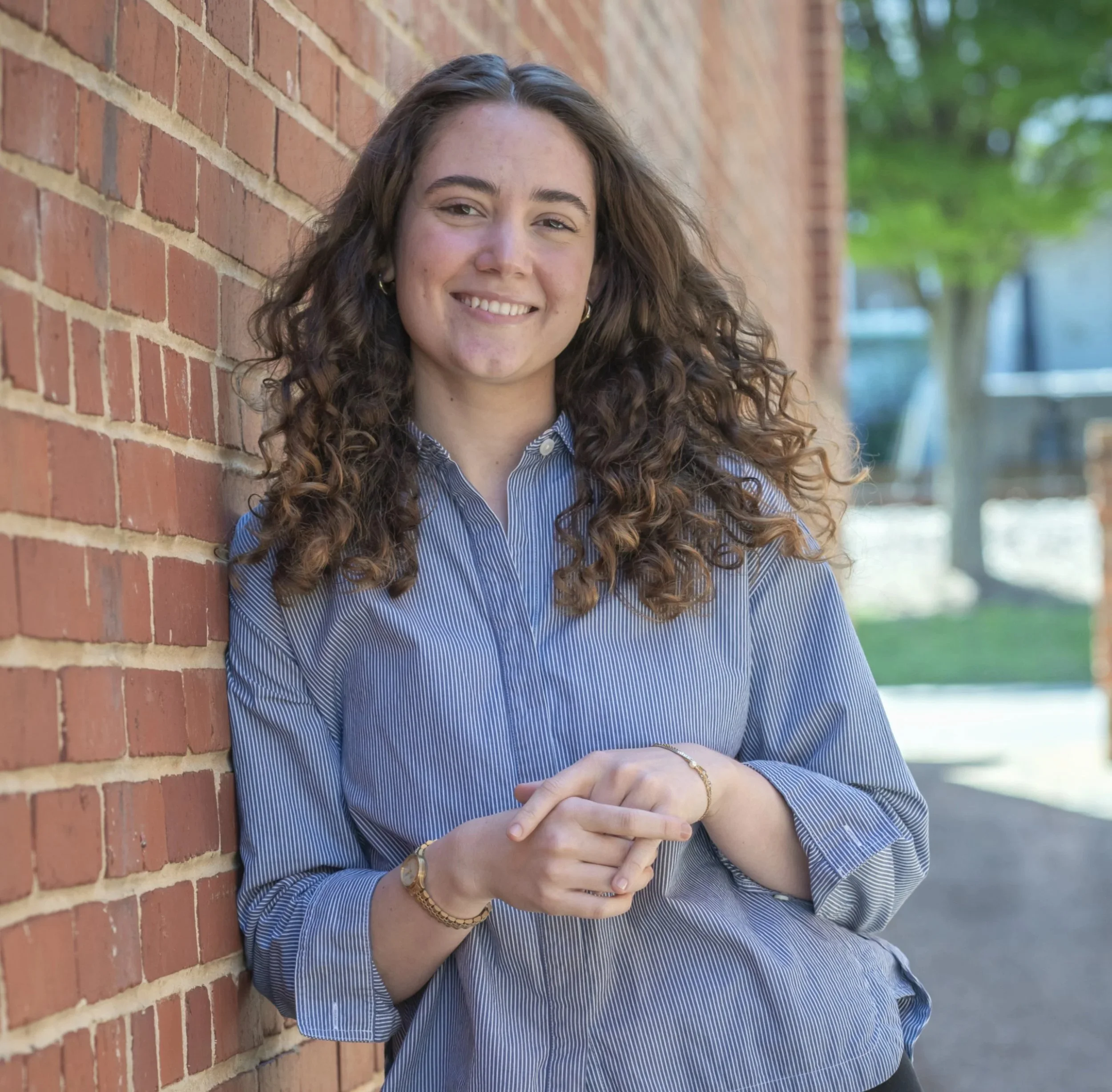 A young woman with curly hair and a big smile leaning against a brick wall outdoors, wearing a blue and white striped shirt, with green trees in the background.