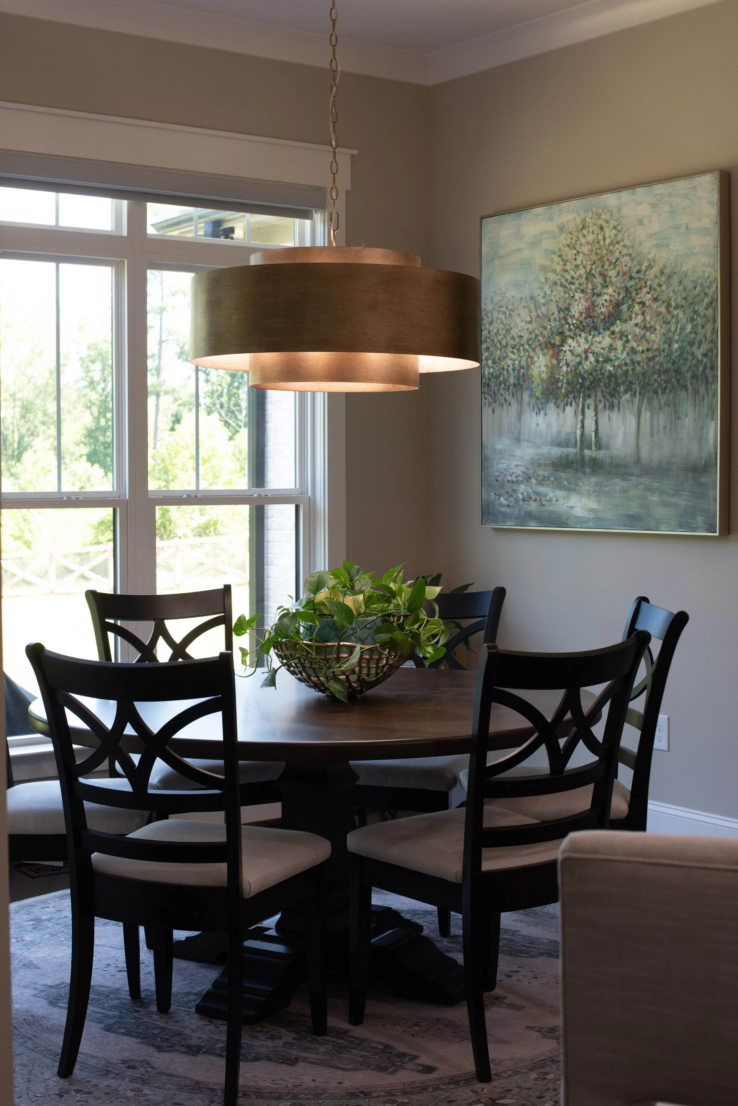 A dining room with a dark wood round table surrounded by six black chairs with beige upholstered seats. A decorative green plant in a woven basket is in the center of the table. Behind the table, there's a large window with grid panes showing greenery outside. To the right, there's a large painting of a tree by a water body hanging on a beige wall. A circular pendant light with a tan fabric shade hangs over the table, centered in the room.