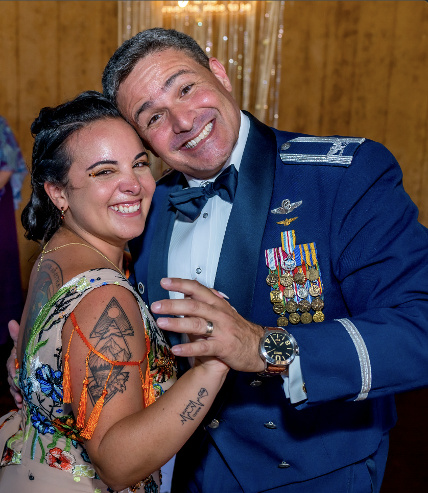 Lisette and John are smiling and dancing at Lisette's father-daughter dance at her wedding. Lisette has dark hair, tattoos on her arm, and is wearing a colorful, patterned dress. The man is in a formal military uniform with many medals and ribbons.