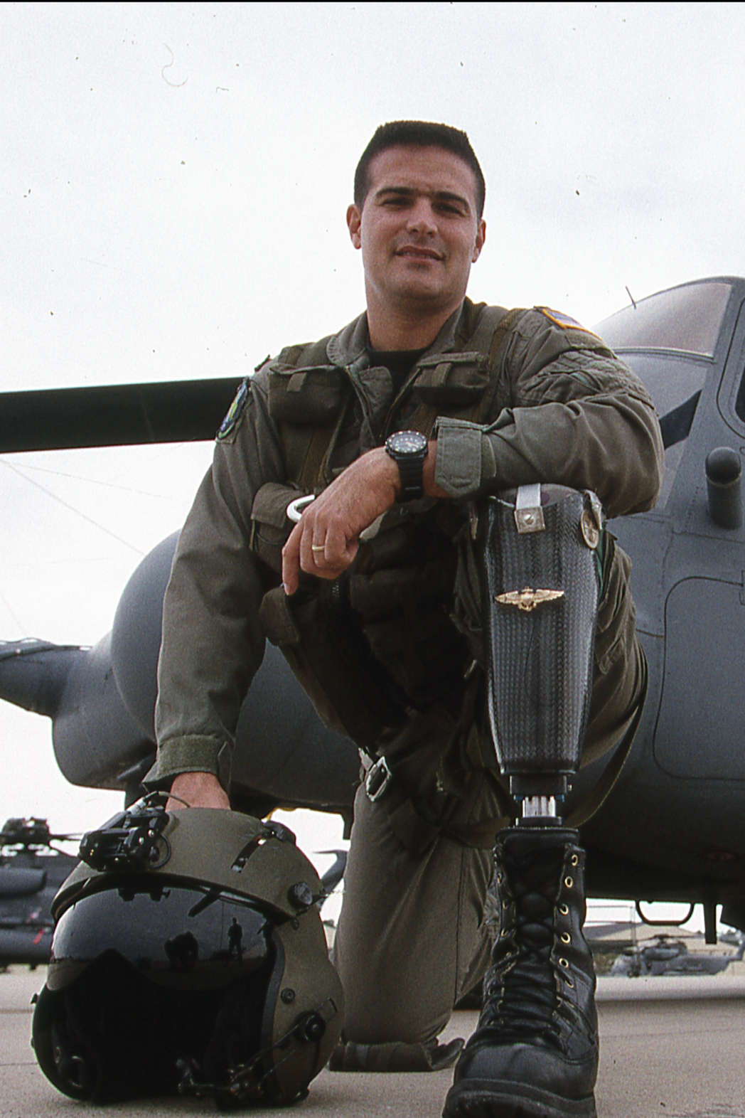 A male soldier in military uniform kneeling in front of a helicopter, with a helmet on the ground beside him.