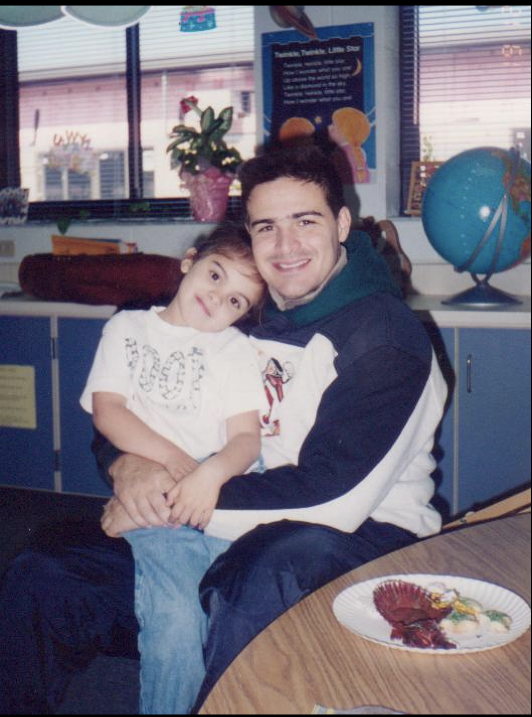A man and a young girl sitting together in a classroom, with the girl leaning on the man's shoulder. The background has a globe, a flower in a pot, and a poster about planets and constellations. A paper plate with a decorated cake or food is on the table in front of them.
