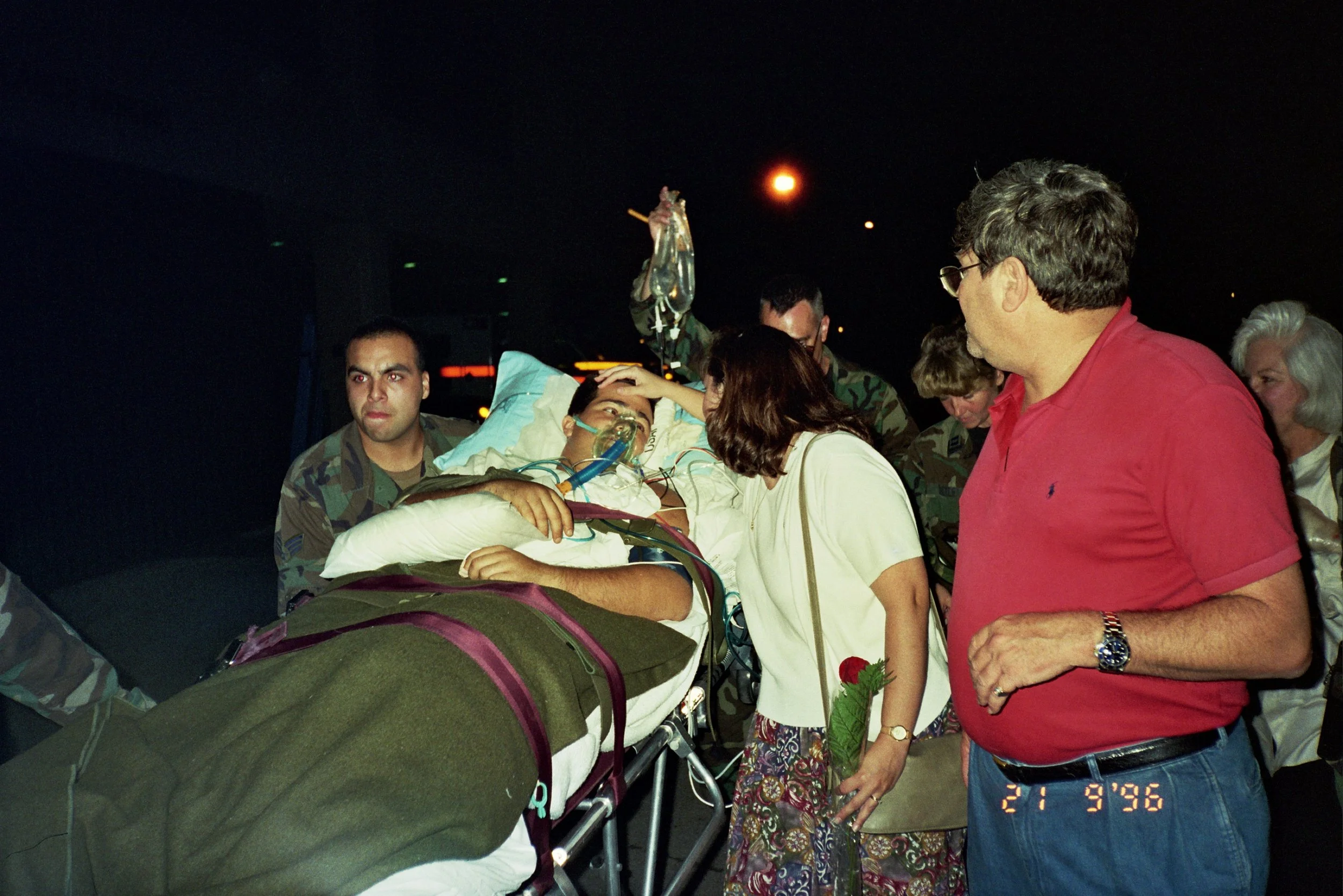 A person in a hospital bed connected to medical equipment, surrounded by military personnel and civilians, with a woman touching the patient's forehead in an emotional moment at night.