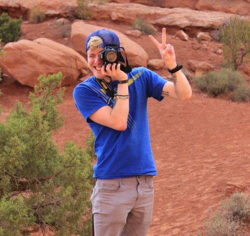 Person wearing blue shirt and cap holding a camera, making a peace sign, standing in a desert landscape with red rocks and shrubbery.