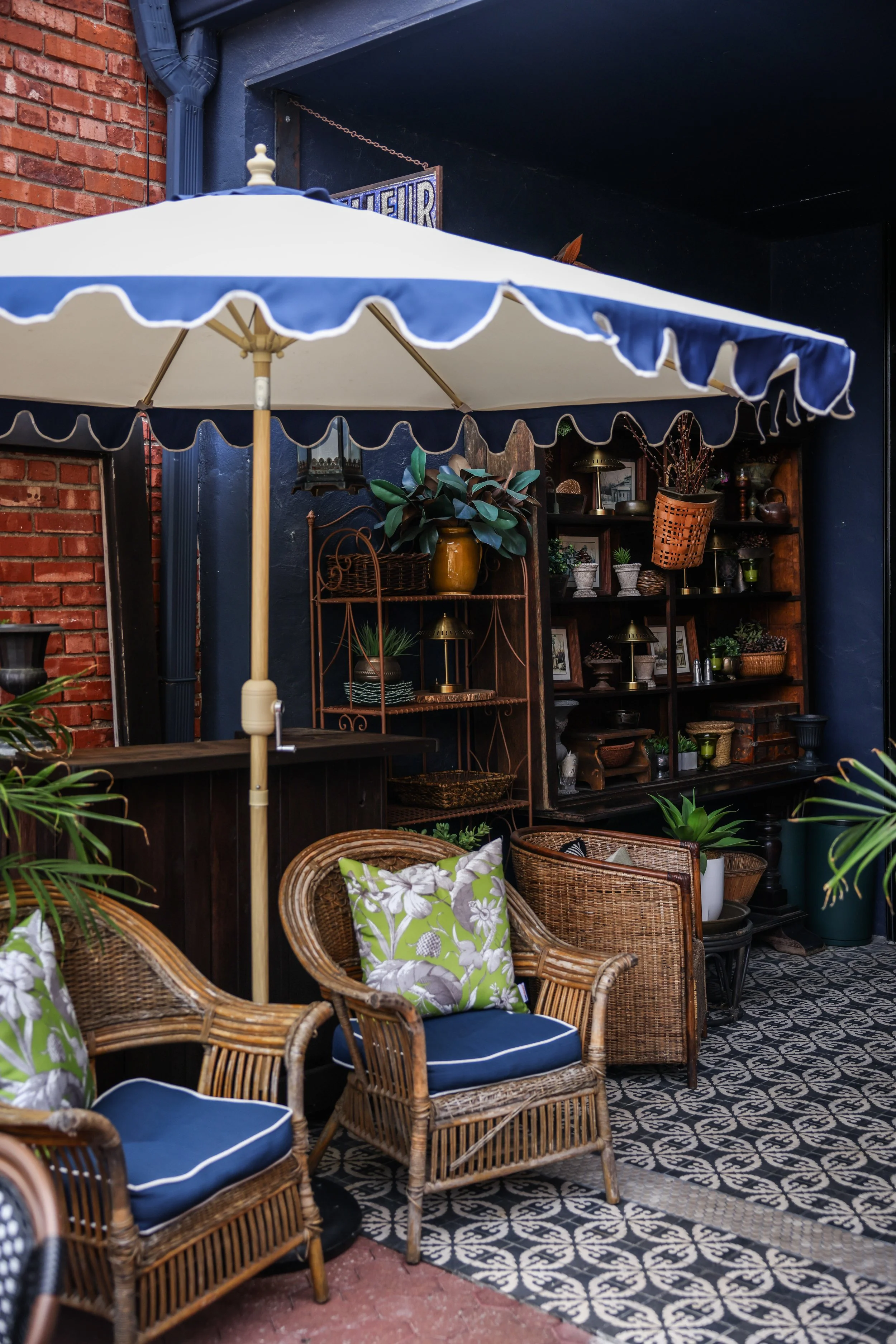 Outdoor patio area with wicker chairs featuring blue cushions and floral pillows, a patterned tile floor, and a dark wooden bookshelf filled with plants, lamps, and decorative items, shaded by a large white and blue umbrella.