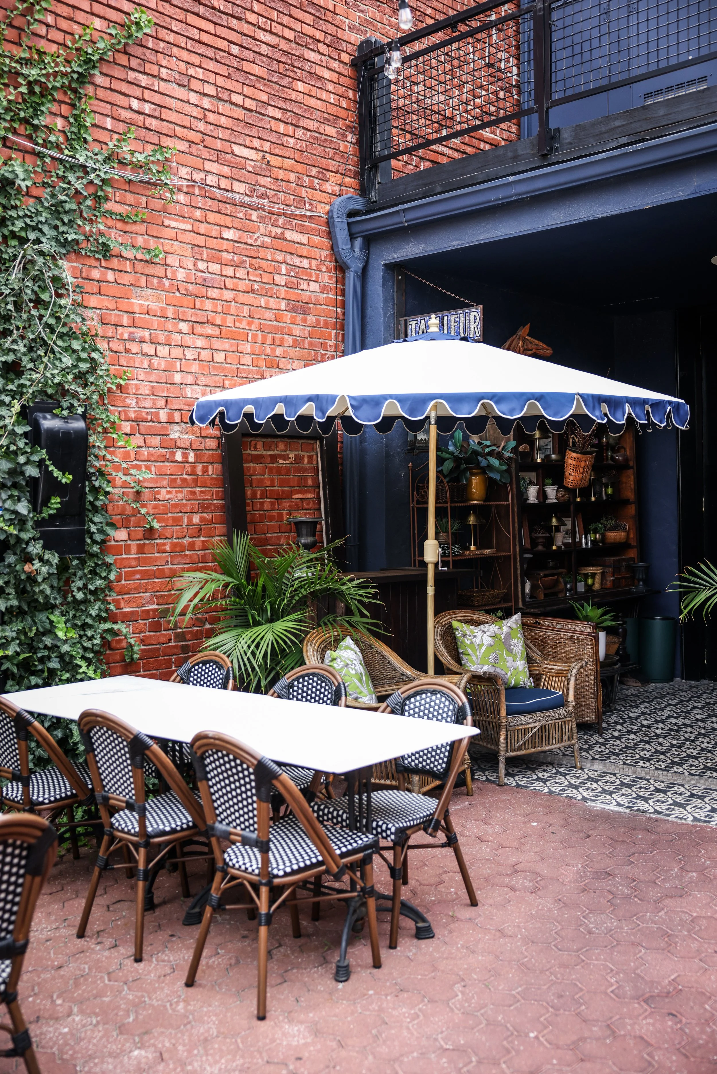 Outdoor patio with tables and chairs, green plants, brick and dark blue walls, and a patio umbrella.
