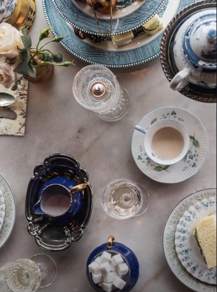 A table set with teacups, a teapot, glassware, a plate with a slice of cake, flowers, and decorative trays.