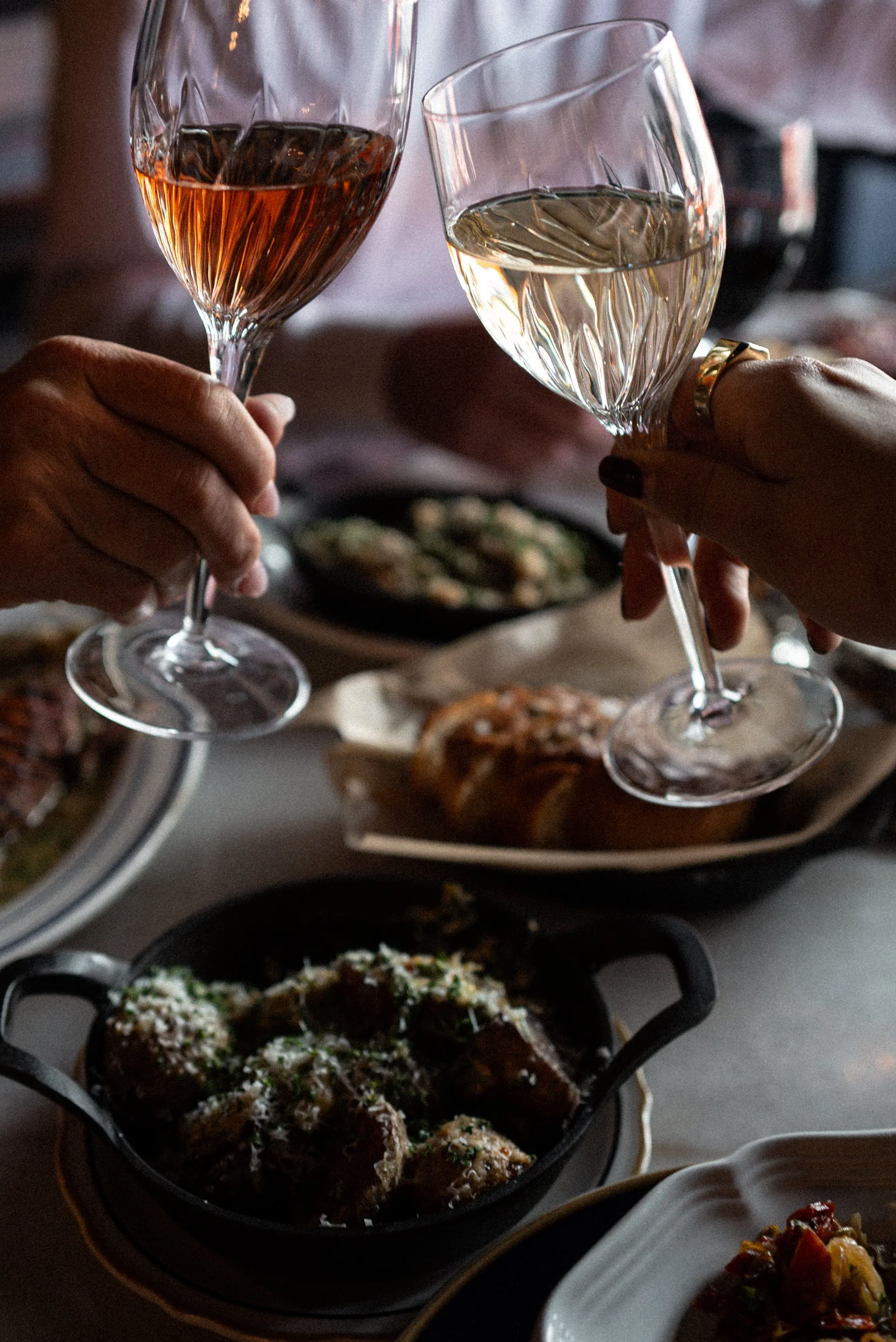 Two people clinking glasses of rosé and white wine above a table with various French inspired dishes, including a cast iron skillet with fresh baked bread, near a casual dining setting.
