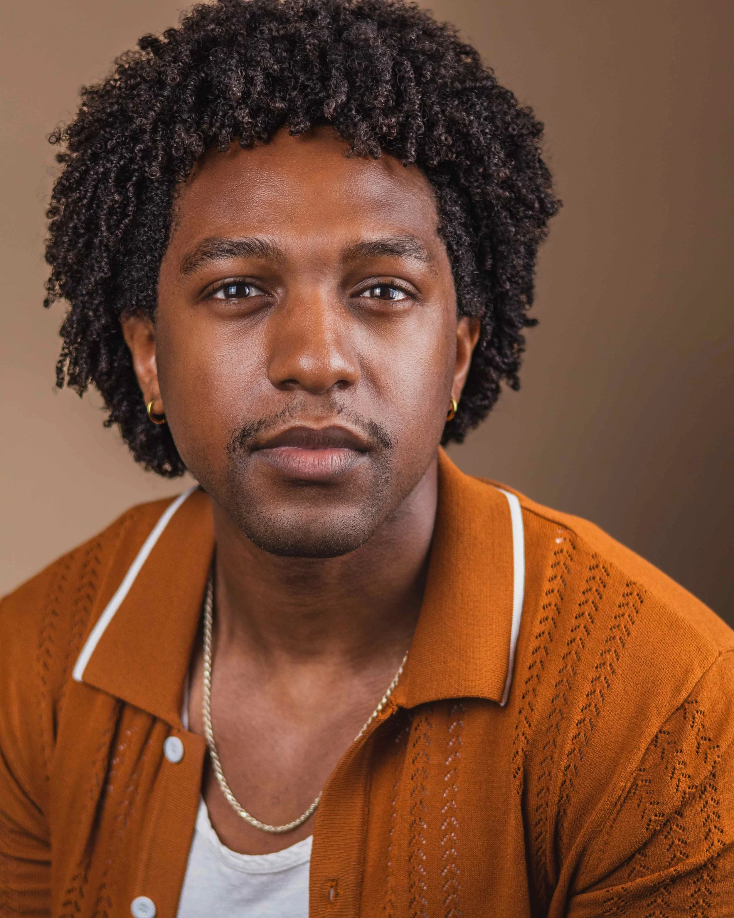Close-up of a man's face and upper torso. He has dark skin, curly black hair, and is wearing a brown knit cardigan over a white shirt, accessorized with a gold chain and earrings. The background is plain and neutral.