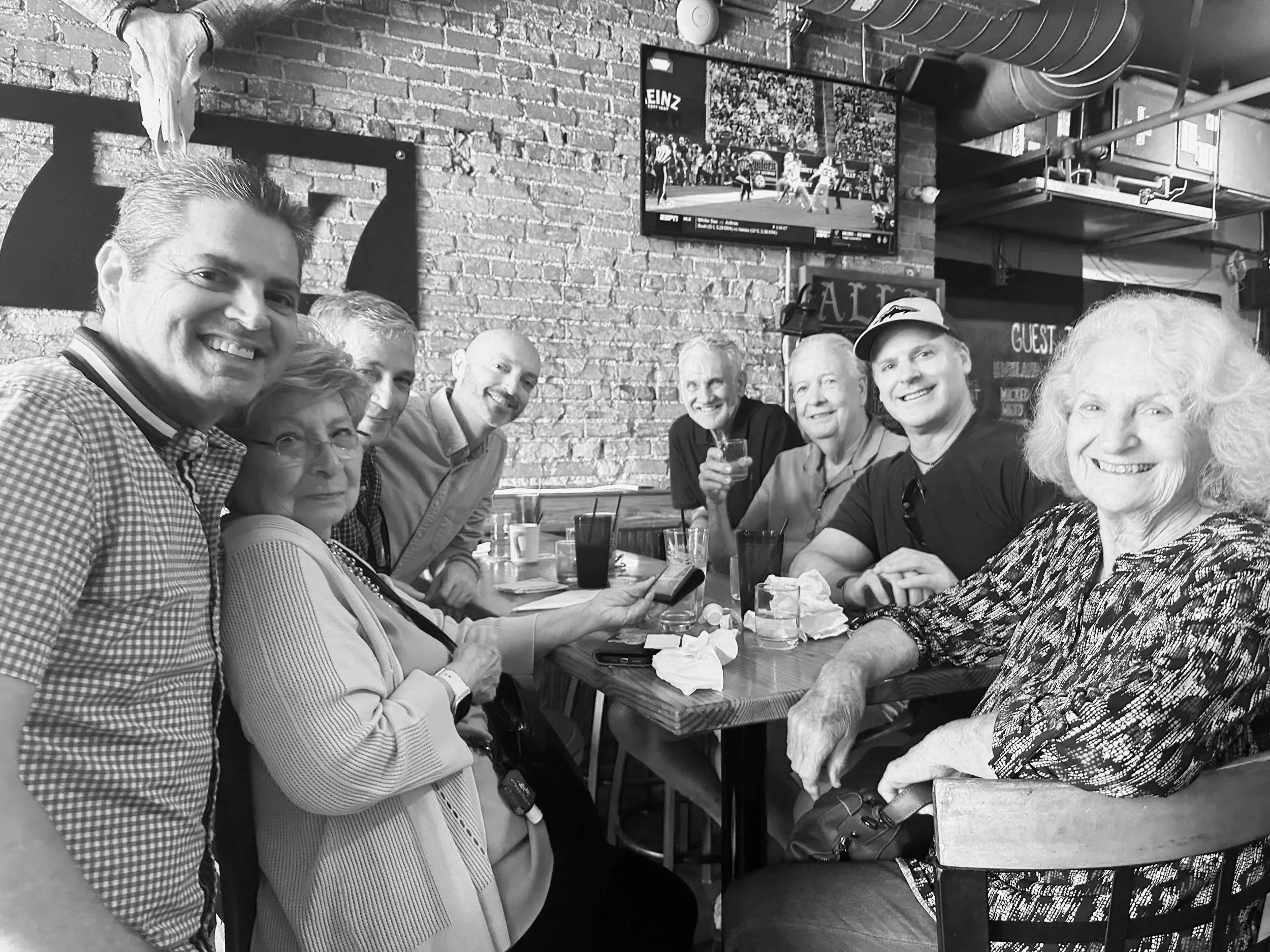 Group of nine people sitting at a table in a restaurant, smiling and looking at the camera, with drinks and napkins on the table, brick wall background, and TV screen on the wall.