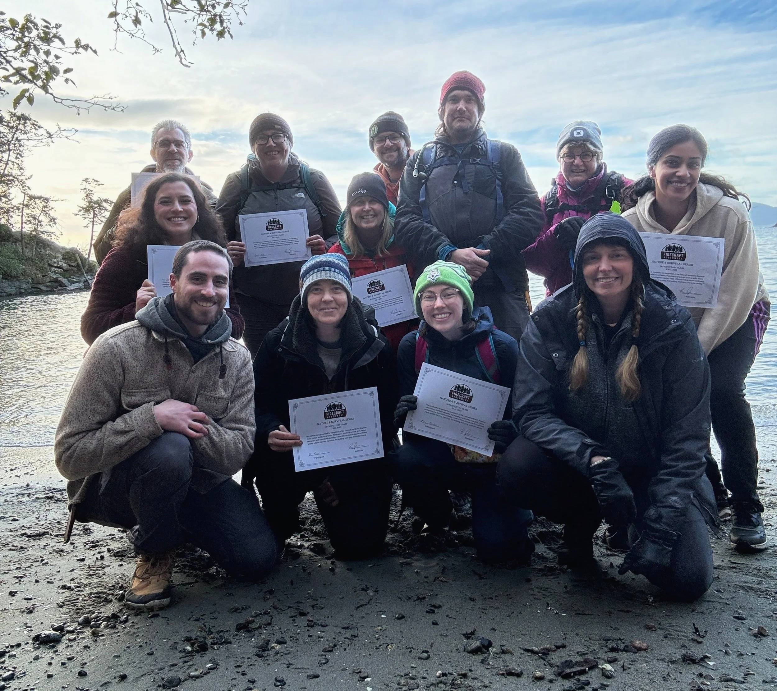 Group of people outdoors on a beach holding certificates, with trees and water in the background.