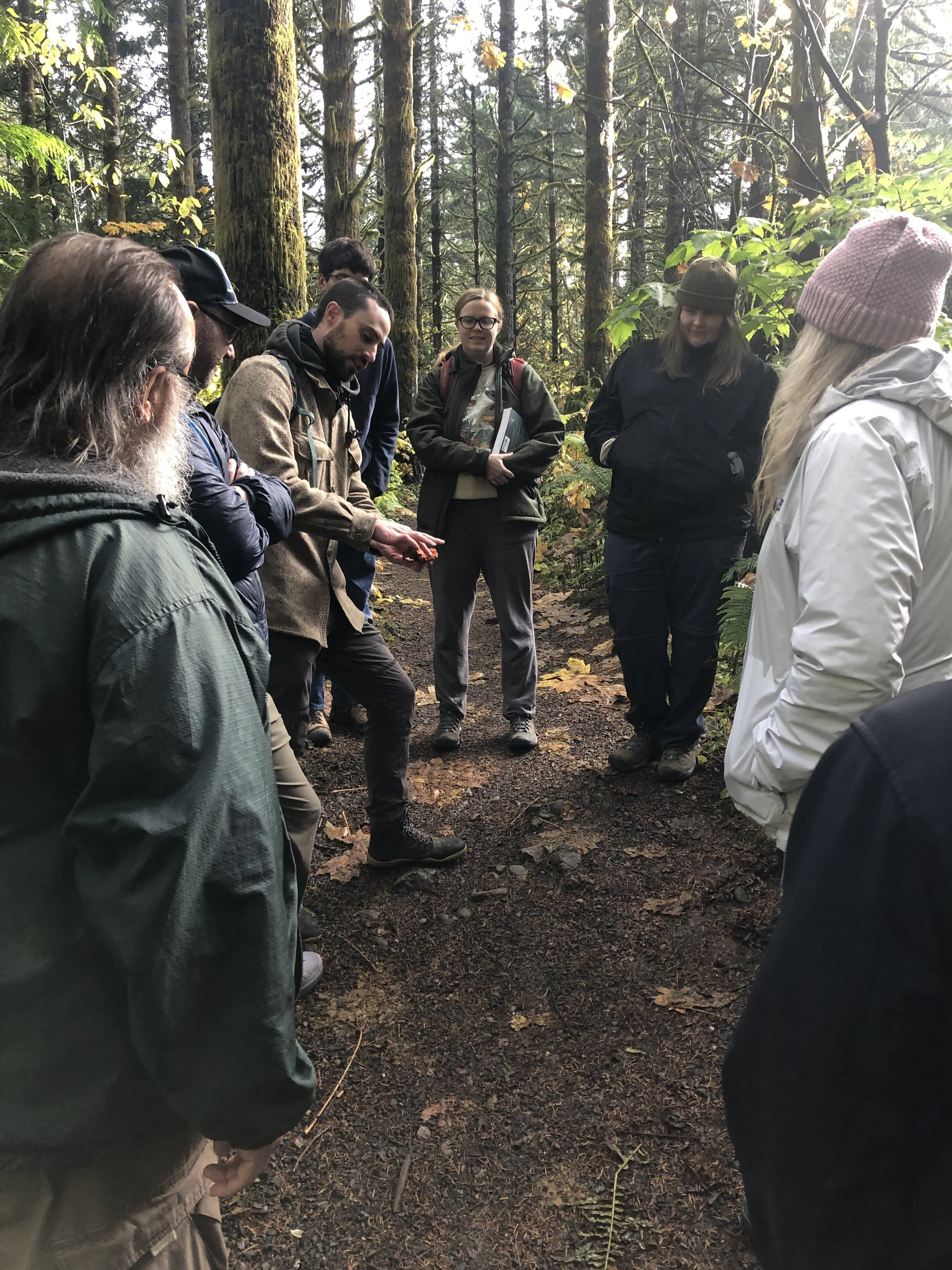 Group of people standing in a forest, one person examining an object, surrounded by trees and foliage.