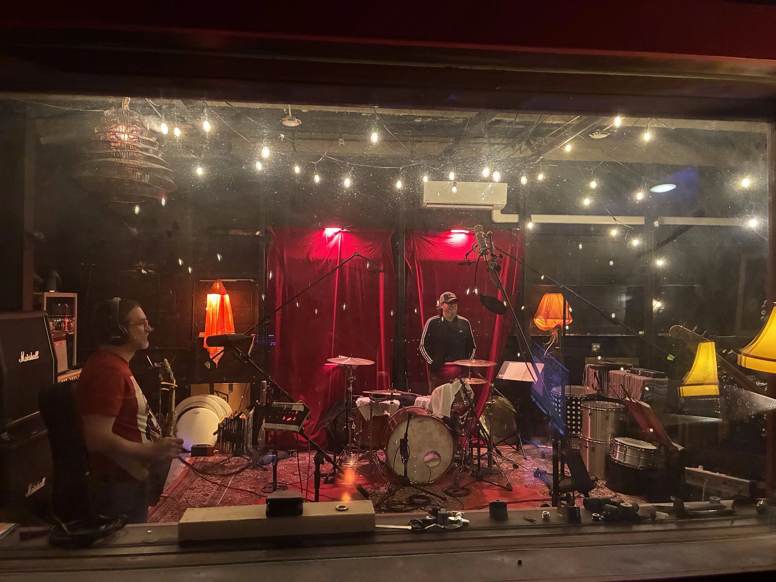 A music rehearsal studio with a band setup, including drums, microphones, and amplifiers, under hanging string lights and a red velvet curtain backdrop. Several people are present, with some seated and others standing, engaged in the musical session.