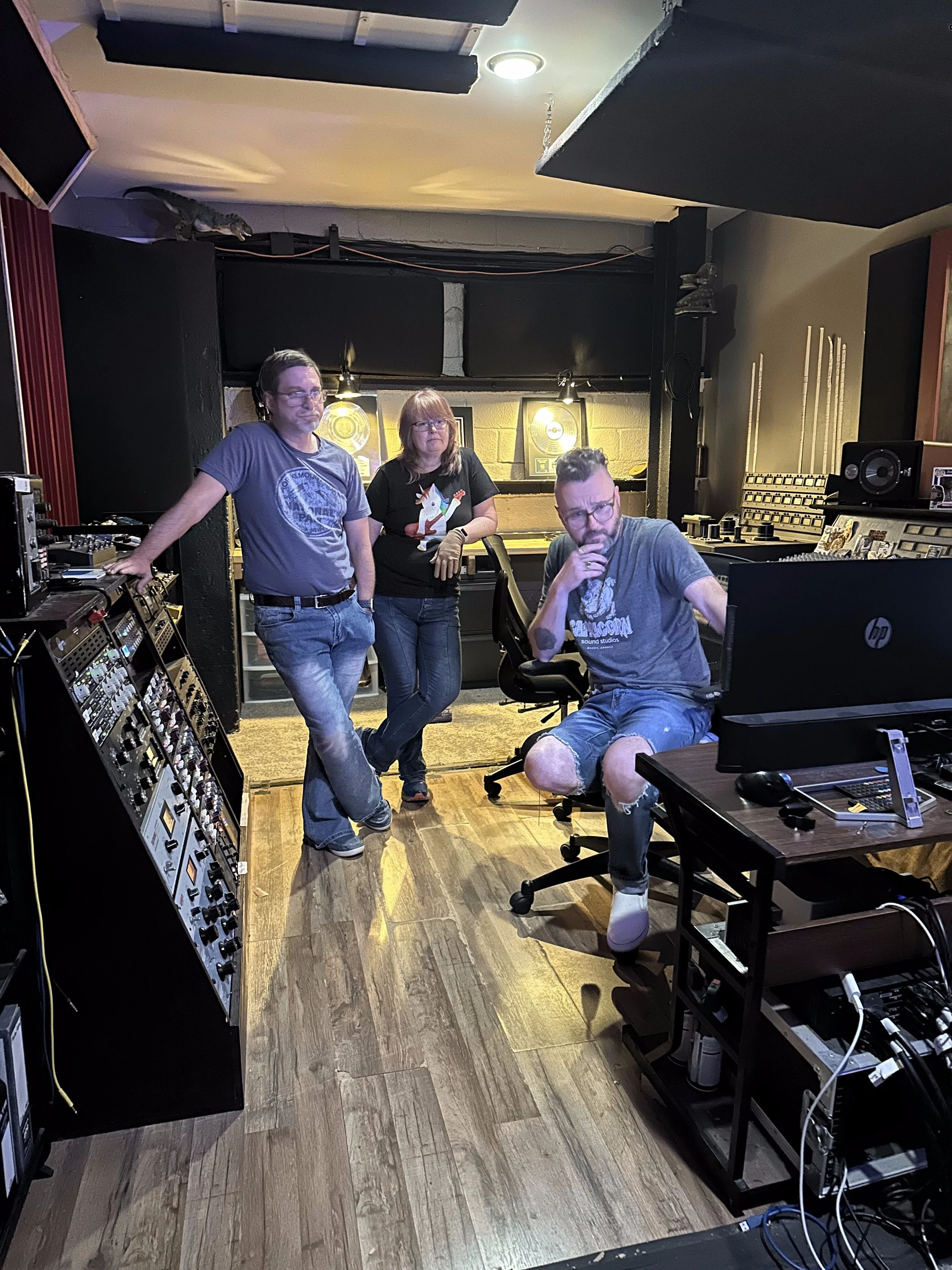 Three people in a music studio surrounded by sound equipment; one seated at a computer and two standing behind, one with hand on hip, the other with arms crossed.