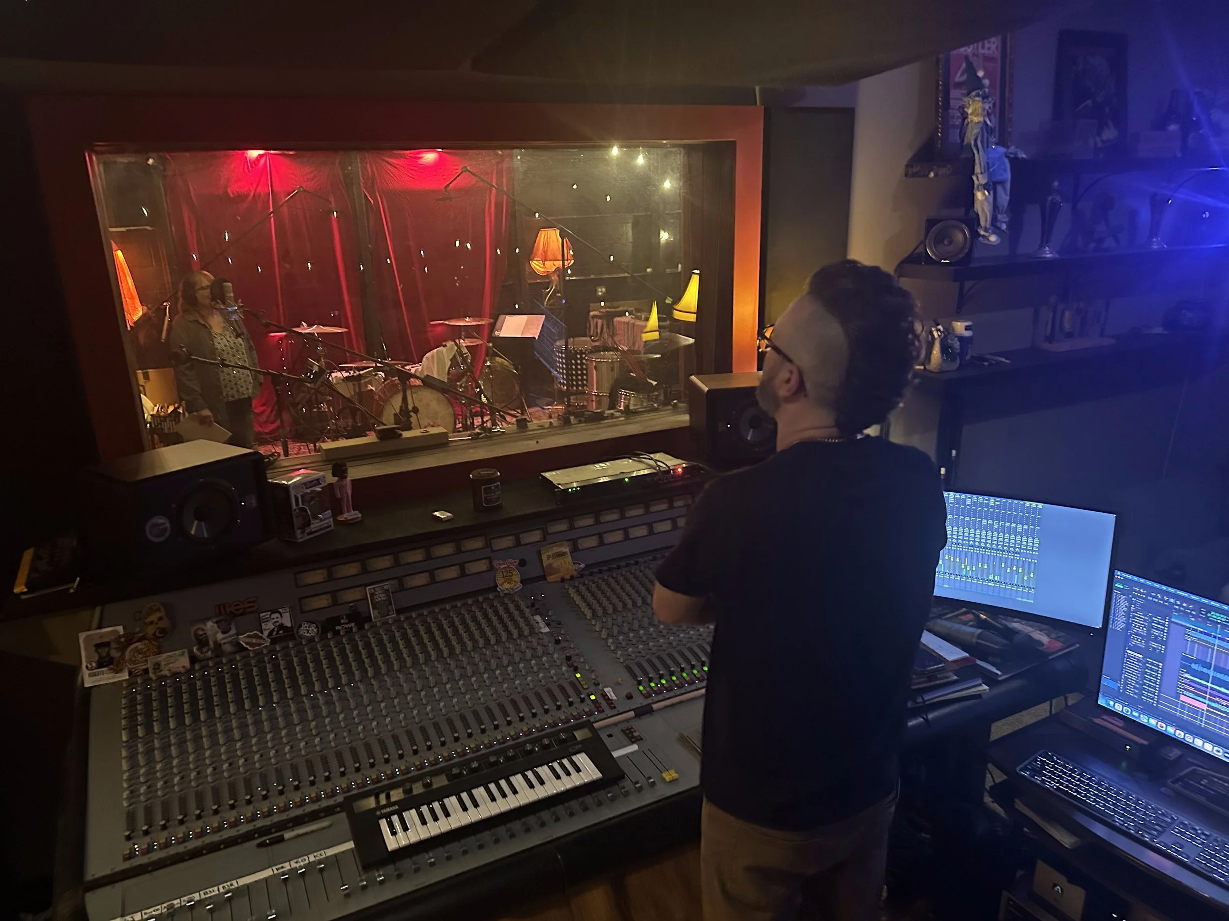 Music recording studio with a person at a mixing console, view through a glass window showing a band performing with drums, microphones, and lamp lighting.