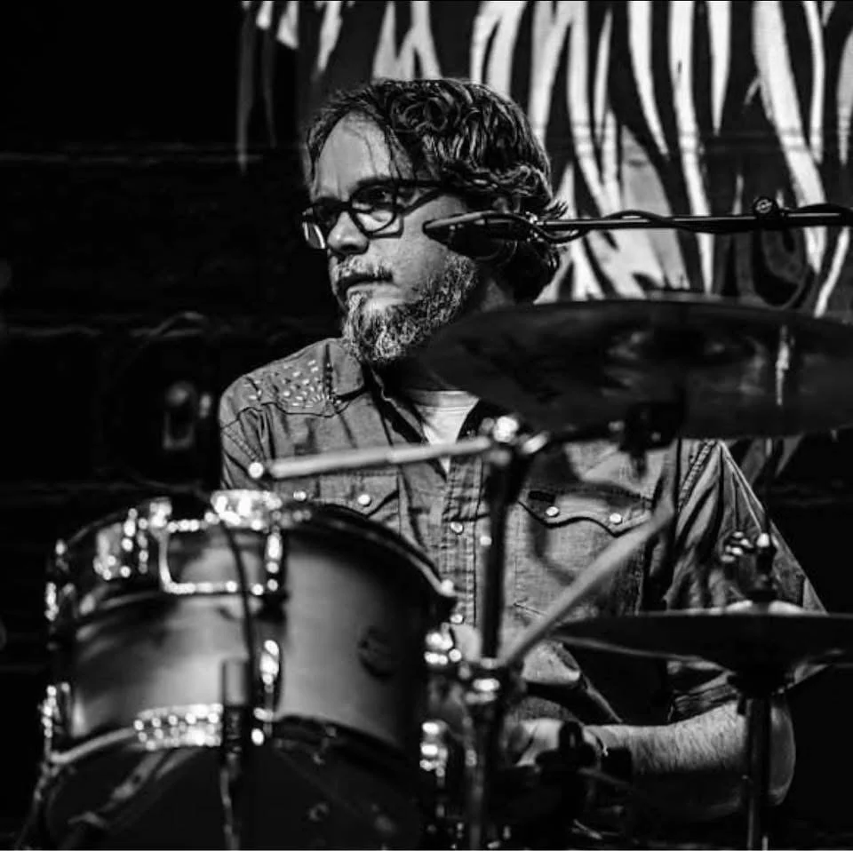 A man with glasses and a beard sits behind a drum set, focused, with a dark, patterned background.