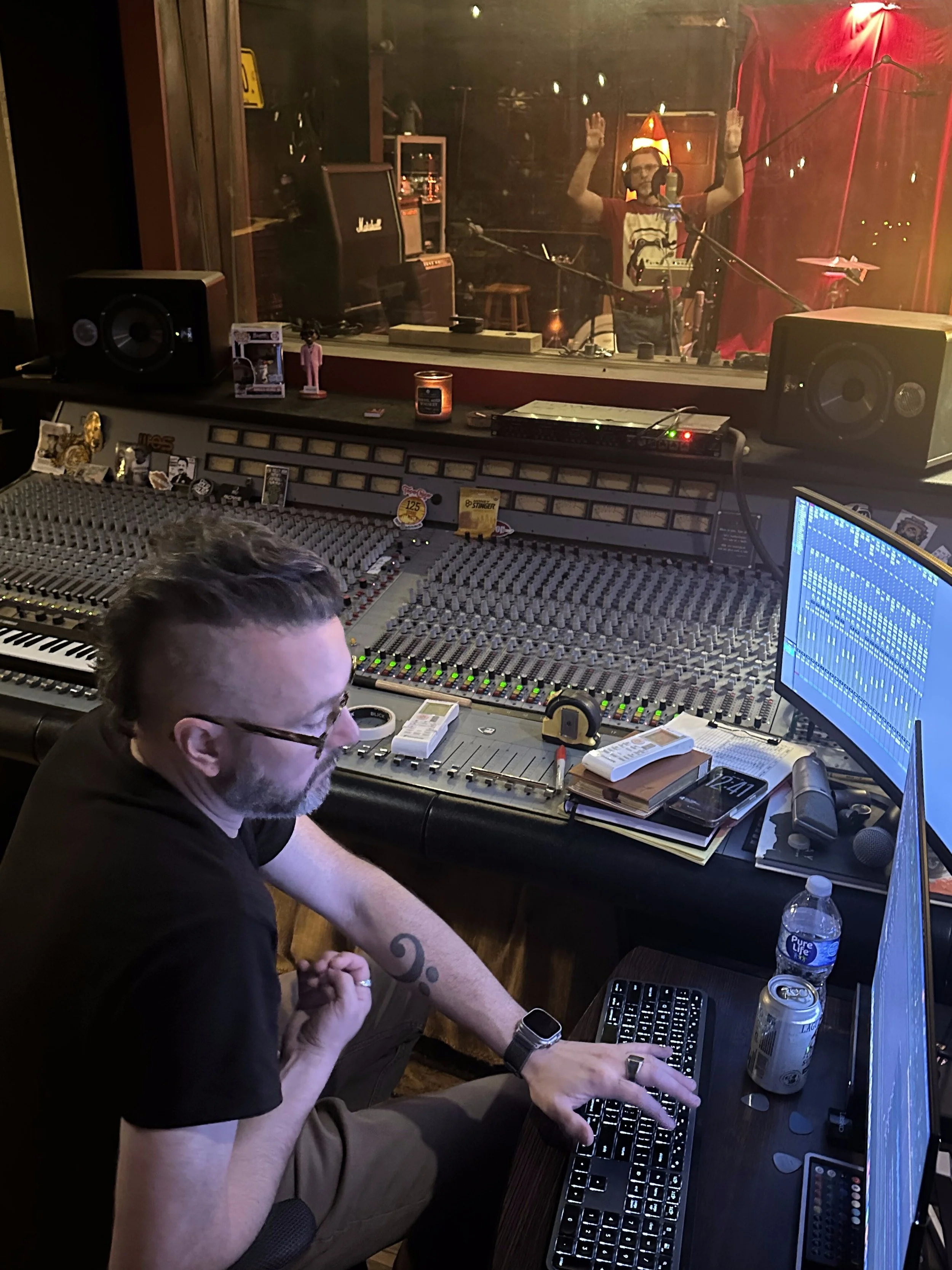 A man with glasses and a beard working in a recording studio, sitting in front of a mixing console with a computer monitor, with various equipment, water bottle, and cans on the desk.