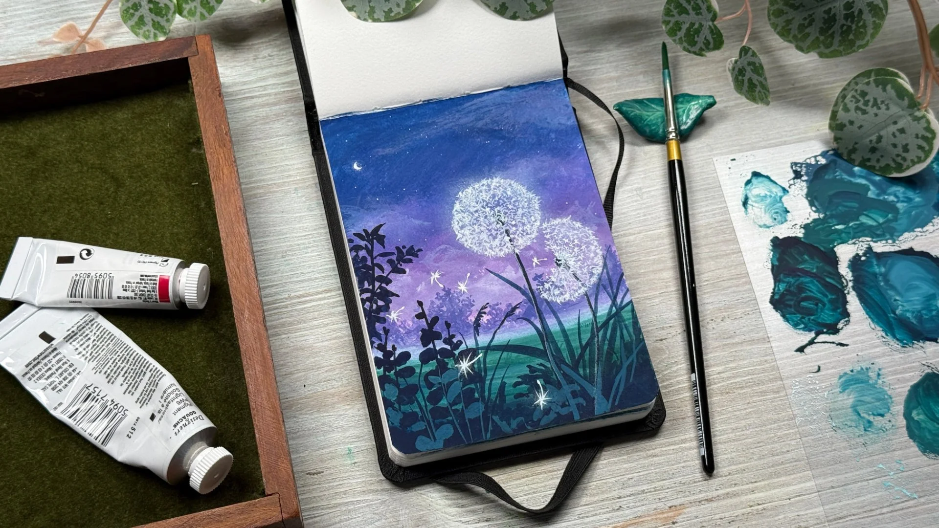 Gouache painting of glowing dandelions at night in a sketchbook, with paint palette and brushes nearby