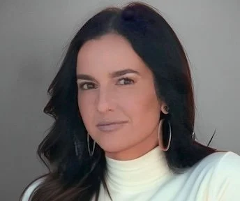 Portrait of a woman with dark hair, wearing a white top and hoop earrings, looking at the camera.