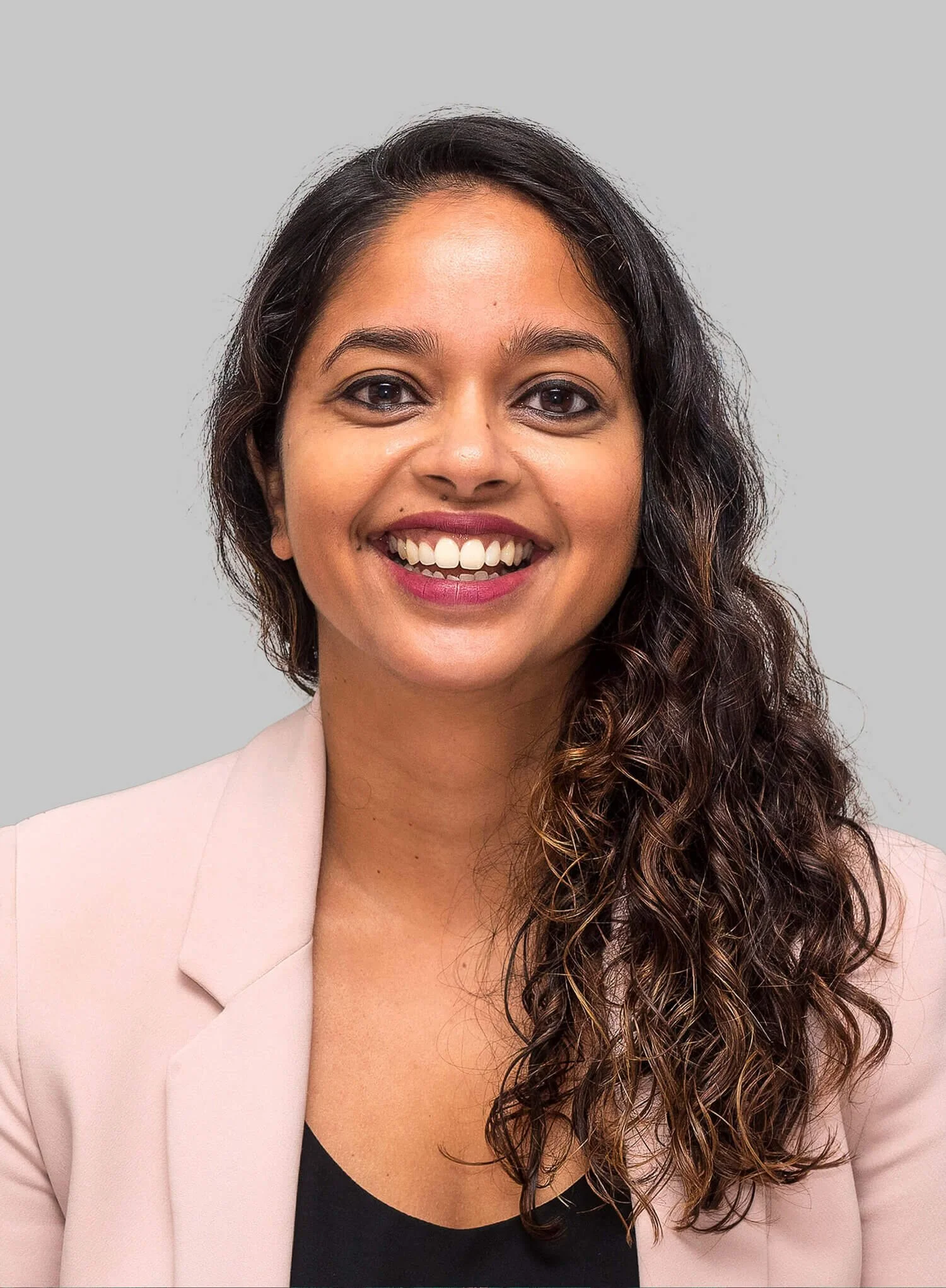 Smiling woman with curly brown hair wearing beige blazer on gray background.