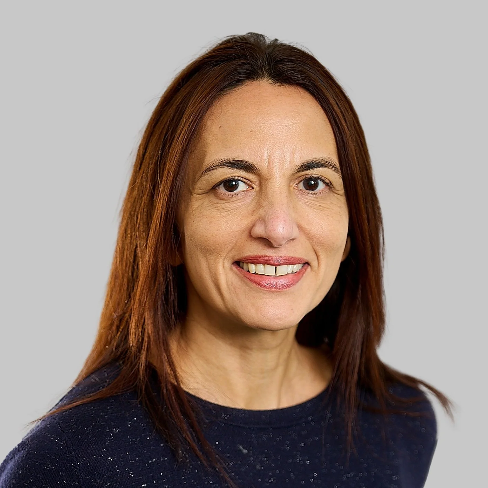 Smiling woman with brown hair wearing navy sparkly top on light gray background.